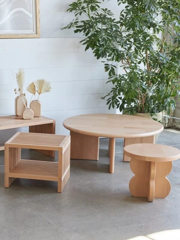 A set of light wood furniture, including a bench, square side table, round coffee table, and small accent table, arranged indoors near a large green plant and wall art.