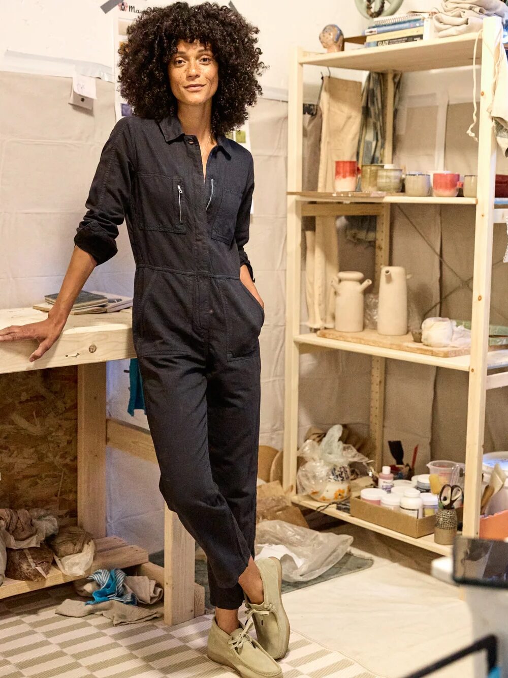 Person in a dark jumpsuit stands in a studio, leaning on a wooden table, with shelves of pottery materials and supplies in the background.