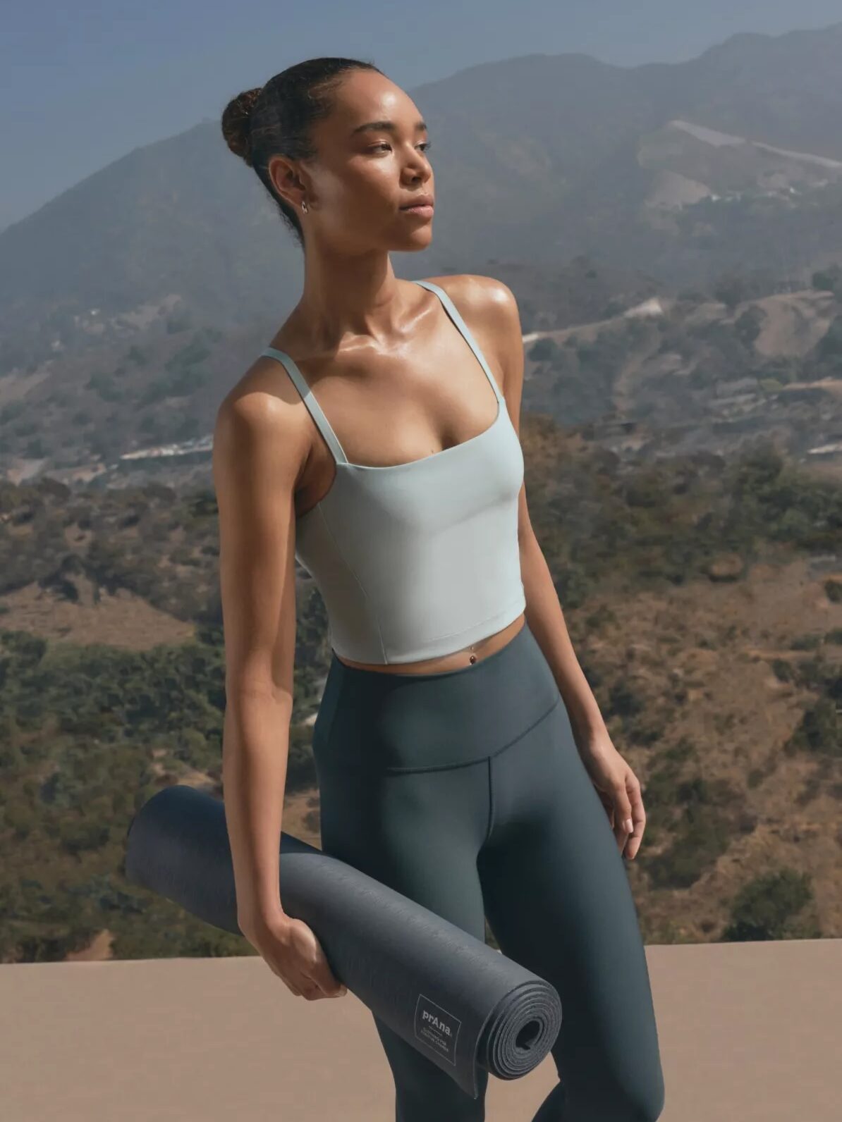 A woman in activewear holds a rolled yoga mat and stands outdoors with mountains and trees in the background.