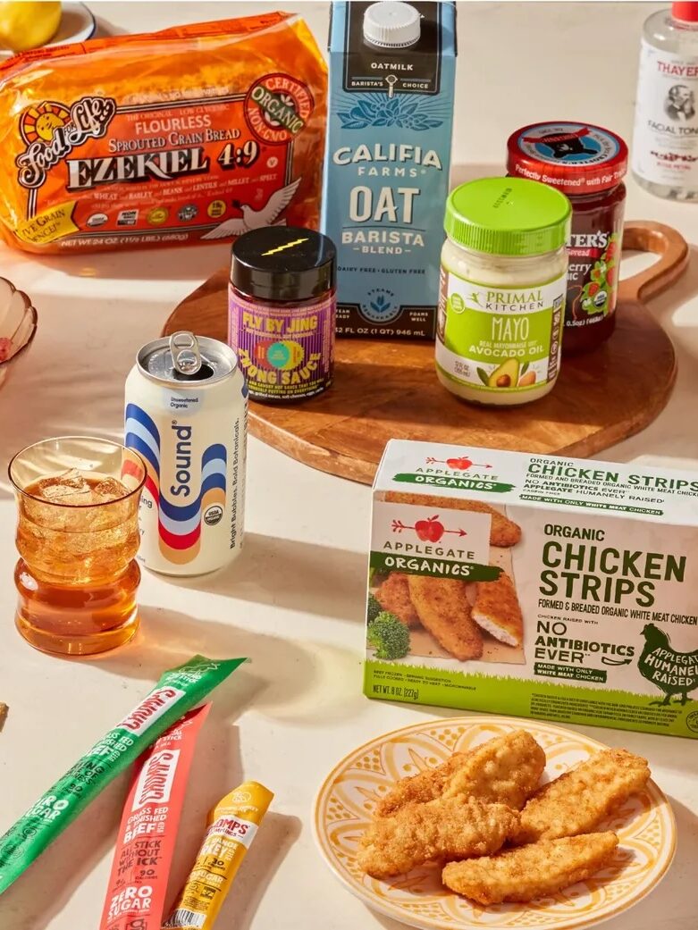 A variety of packaged food products, snacks, drinks, and condiments are displayed on a table with plates, bowls, and glasses.