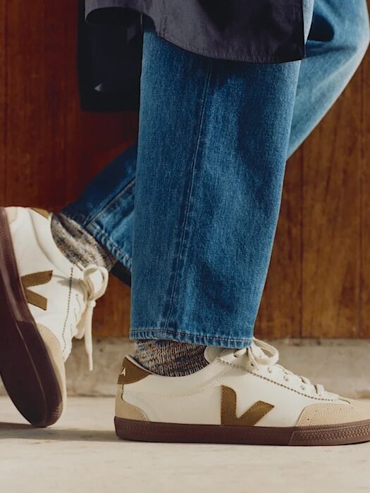 Person wearing blue jeans and white sneakers with beige and brown accents, standing indoors on a light-colored floor near a wooden wall.