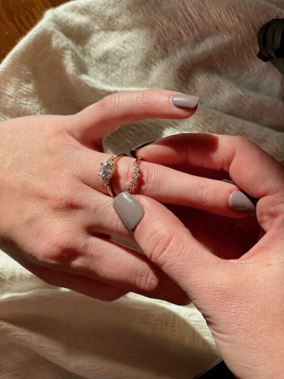 A person places a gold ring with a diamond on another person's finger, both with manicured nails painted gray, on a light-colored fabric background.