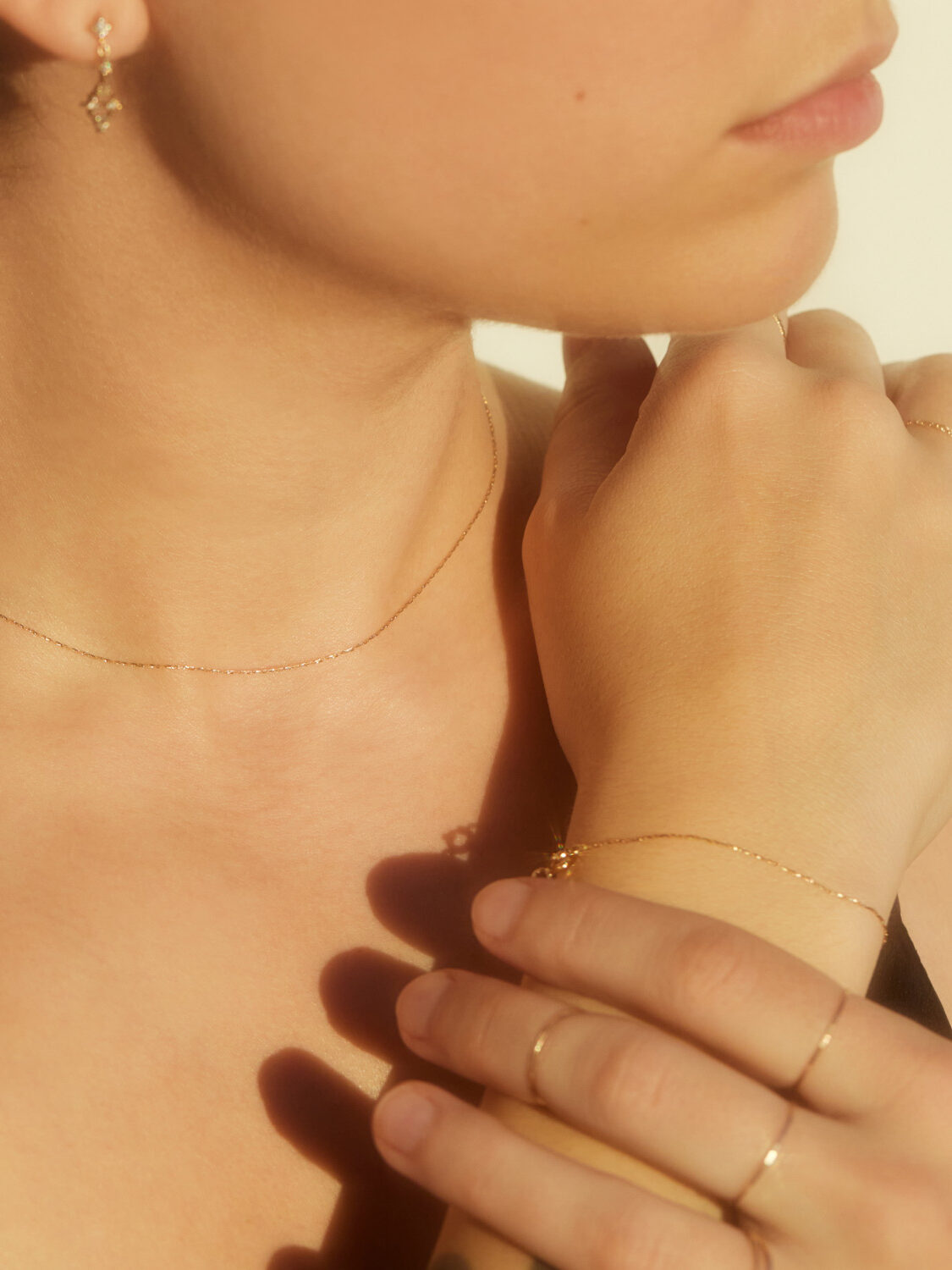 A woman wearing delicate gold jewelry, including a thin necklace, bracelet, earrings, and rings, with soft natural lighting highlighting her skin.
