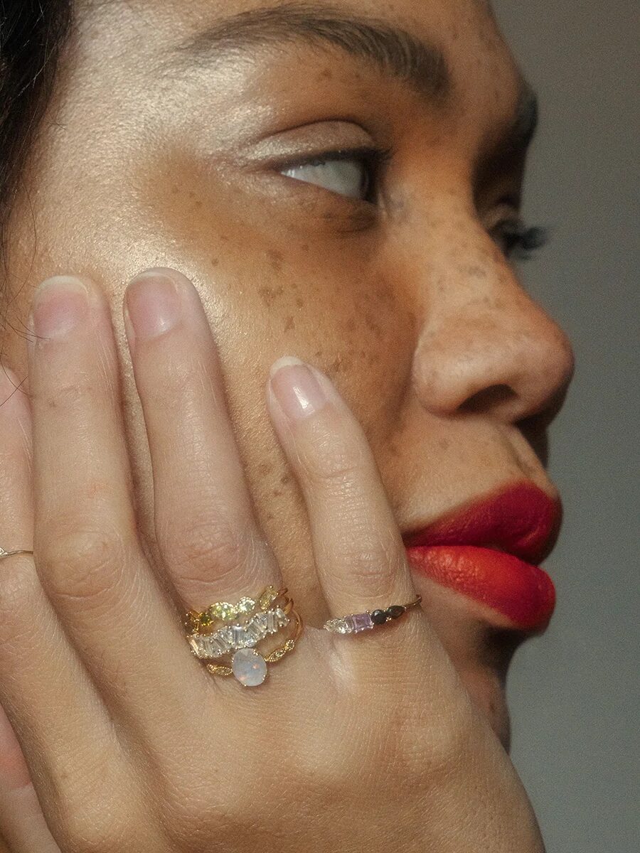 A person with red lipstick and freckles touches their face, displaying multiple gold rings with gemstones on their fingers.