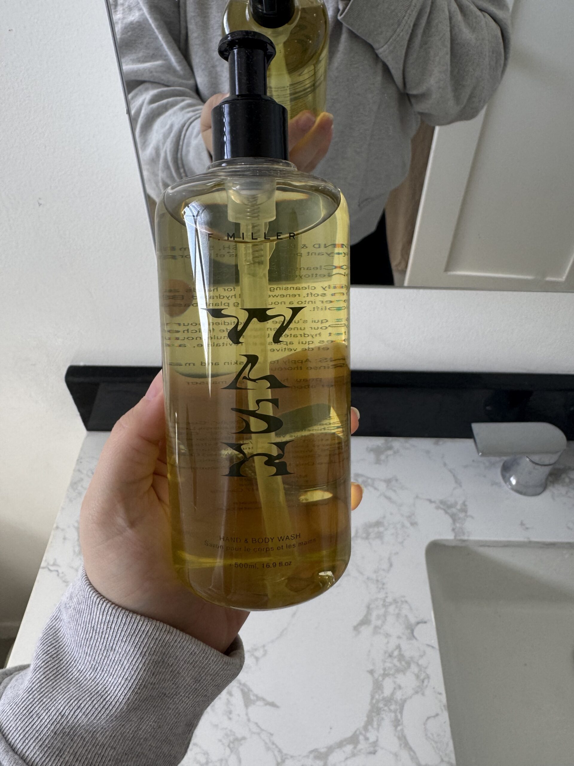 Person holding a bottle of amber body wash with black text and a pump dispenser, standing in front of a bathroom mirror and sink.