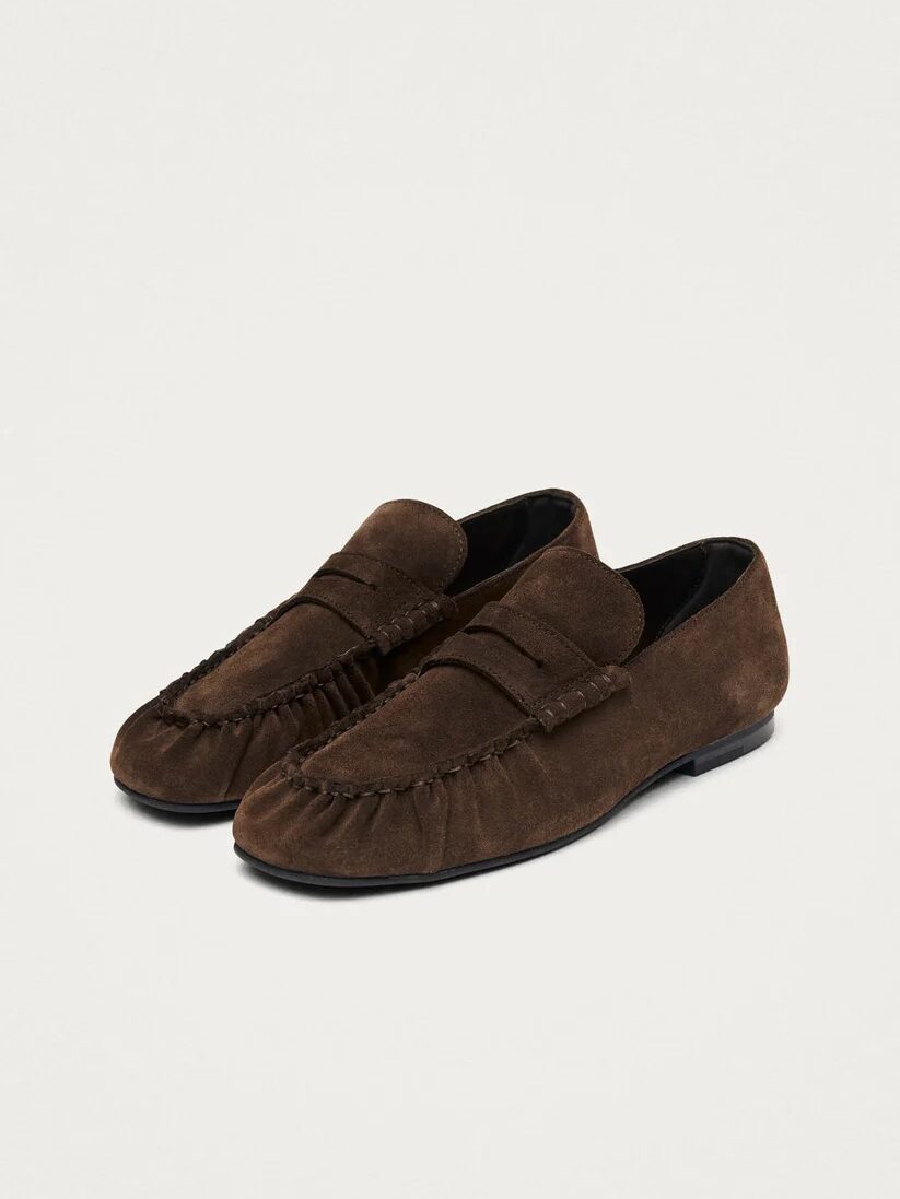 A pair of brown suede loafers with moccasin-style stitching and flat black soles, displayed on a plain light background.