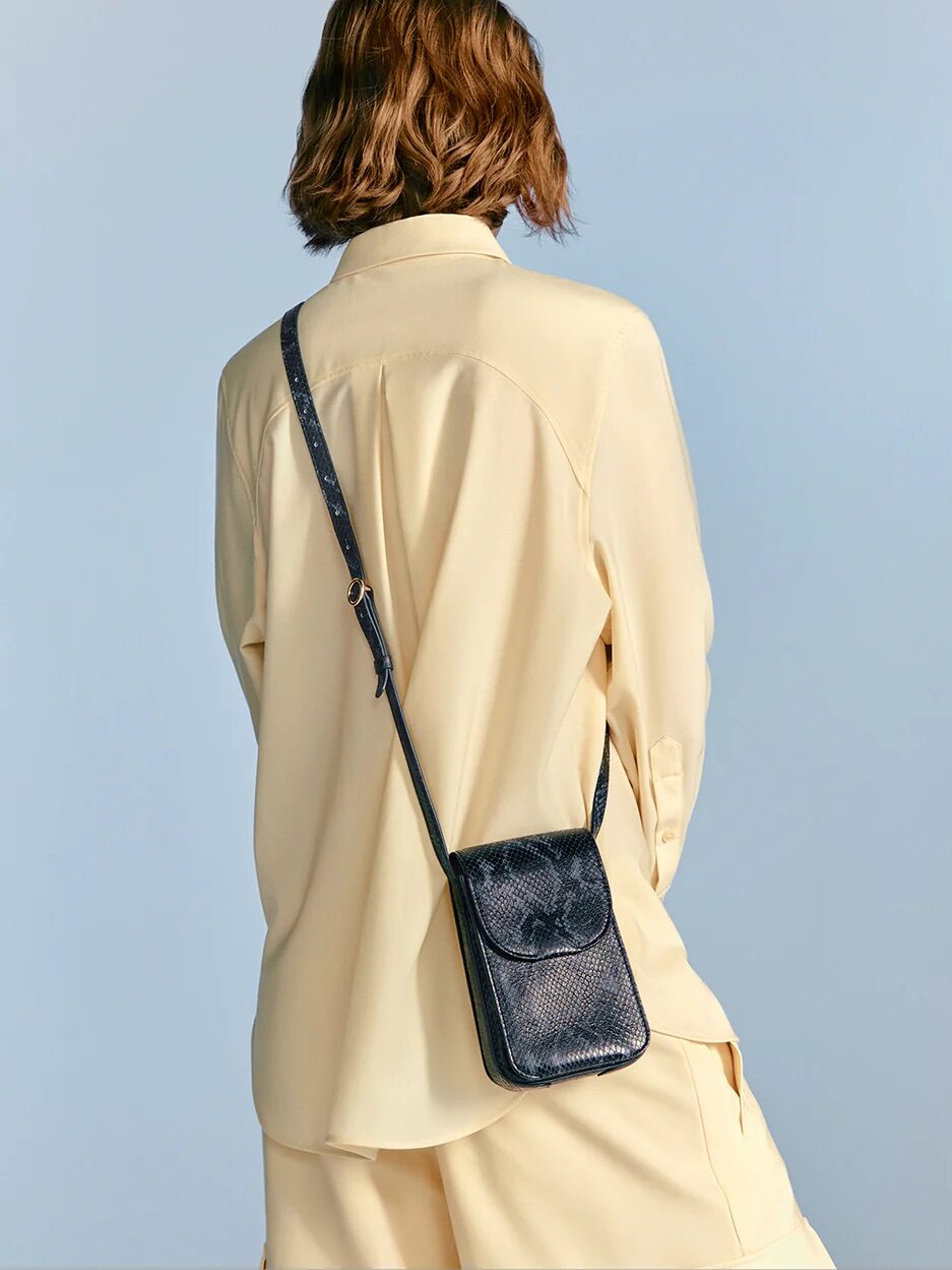 Person with short, wavy hair wears a loose, light beige shirt and shorts, shown from behind, with a black textured crossbody bag over the shoulder.
