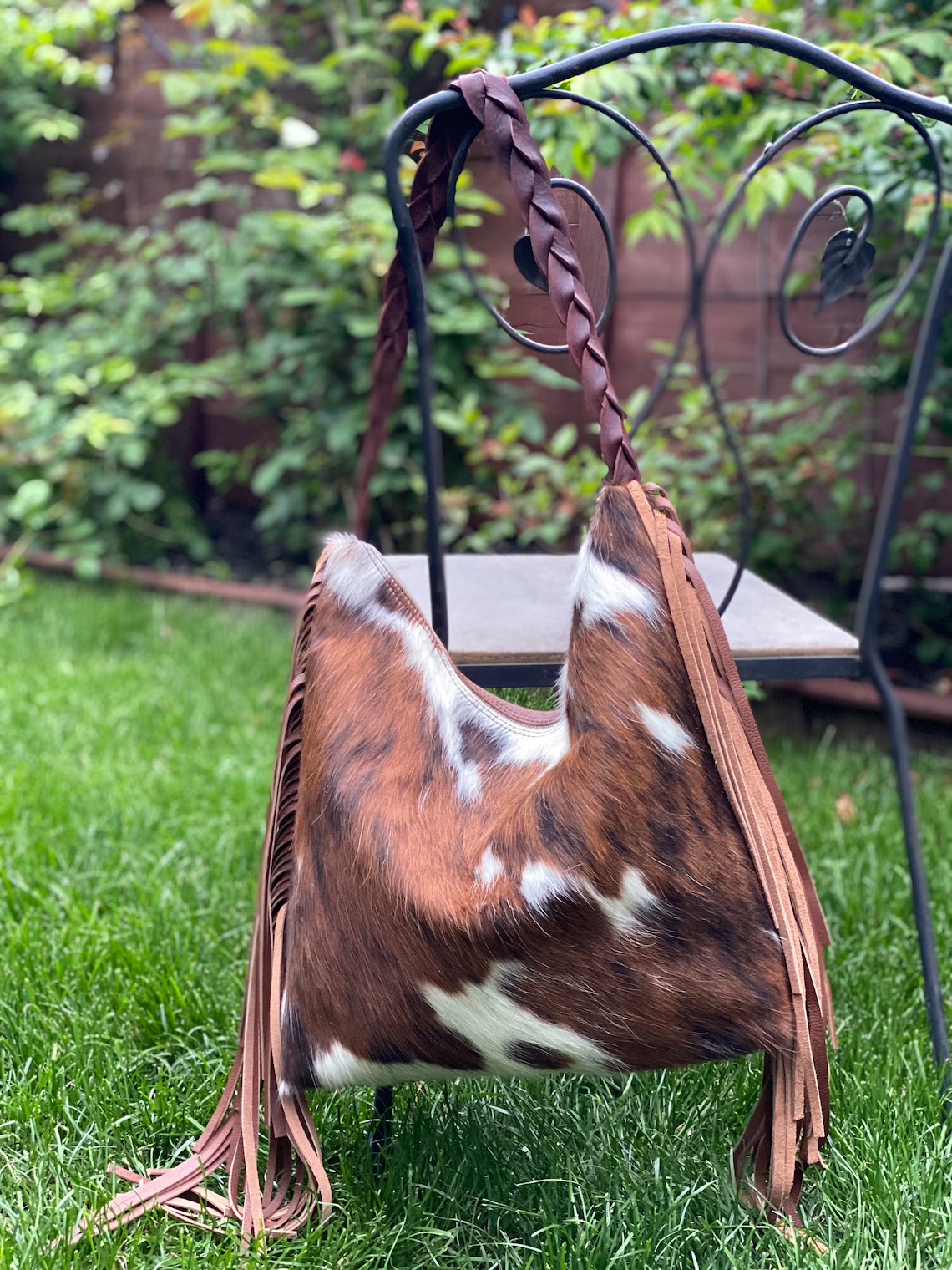 A brown and white cowhide handbag with fringe and a braided strap hangs on a black metal chair in a grassy garden.
