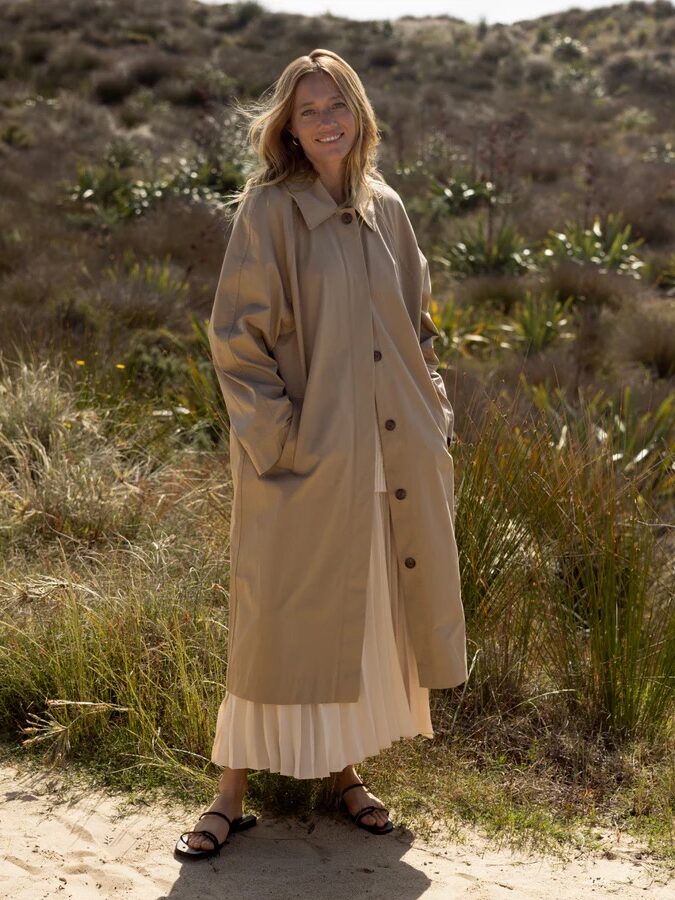 A person stands outdoors on sandy ground, wearing a long beige trench coat, white skirt, and sandals, surrounded by grass and shrubs.