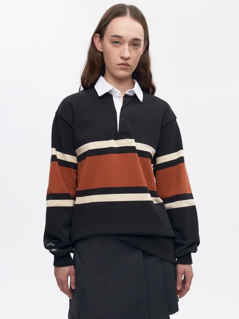 A person wearing a black, orange, and beige striped rugby sweater with a white collar and a dark gray pleated skirt, standing against a plain background.