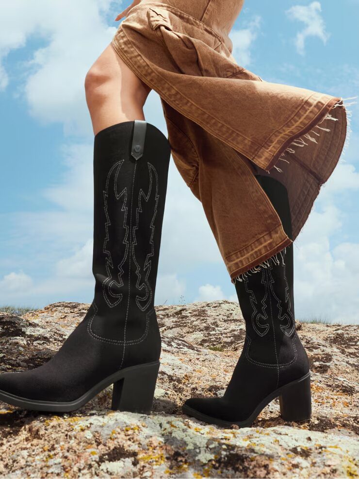 Person wearing a brown skirt and black embroidered heeled cowboy boots standing on rocky ground under a blue sky with clouds.