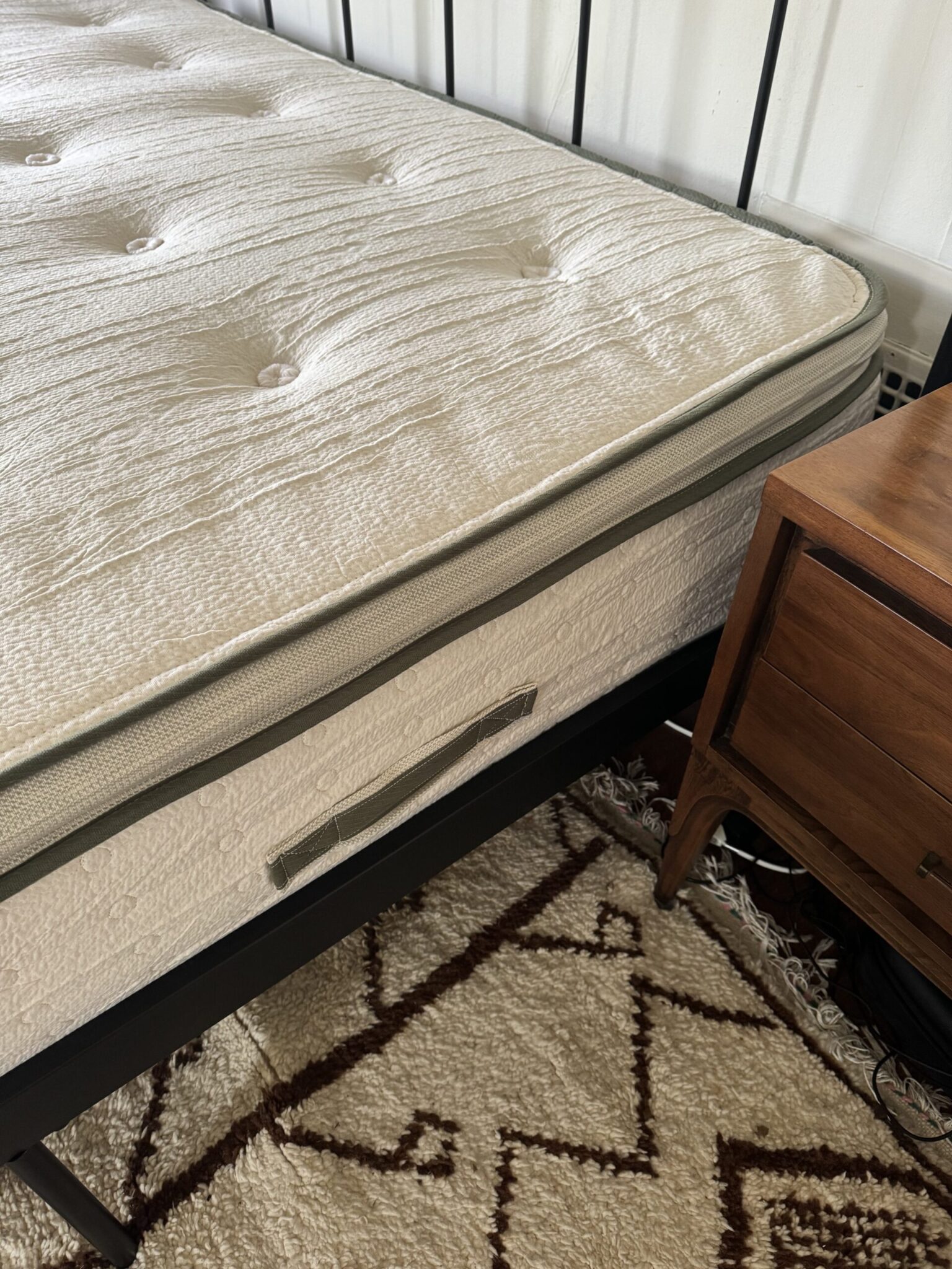 A close-up view of a mattress with a fabric handle, placed on a black metal bed frame next to a wooden nightstand and a patterned rug.