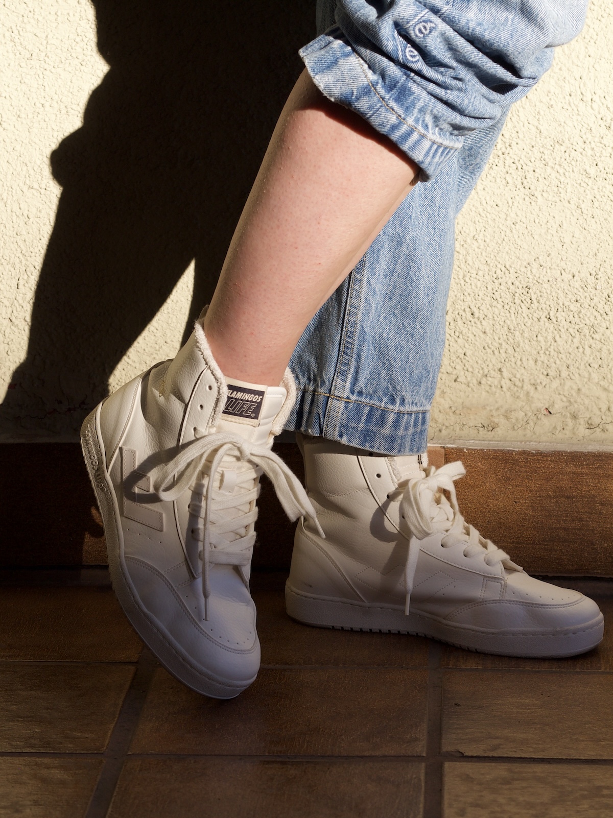 A person wearing light denim jeans with rolled cuffs and sustainable white high-top sneakers stands on a tiled floor near a sunlit wall.