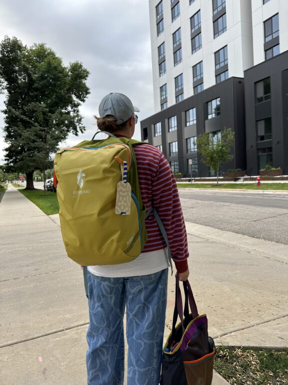 A person wearing a gray cap, striped sweater, and blue patterned jeans stands on a sidewalk near a modern apartment building, carrying a green backpack and a black tote bag.