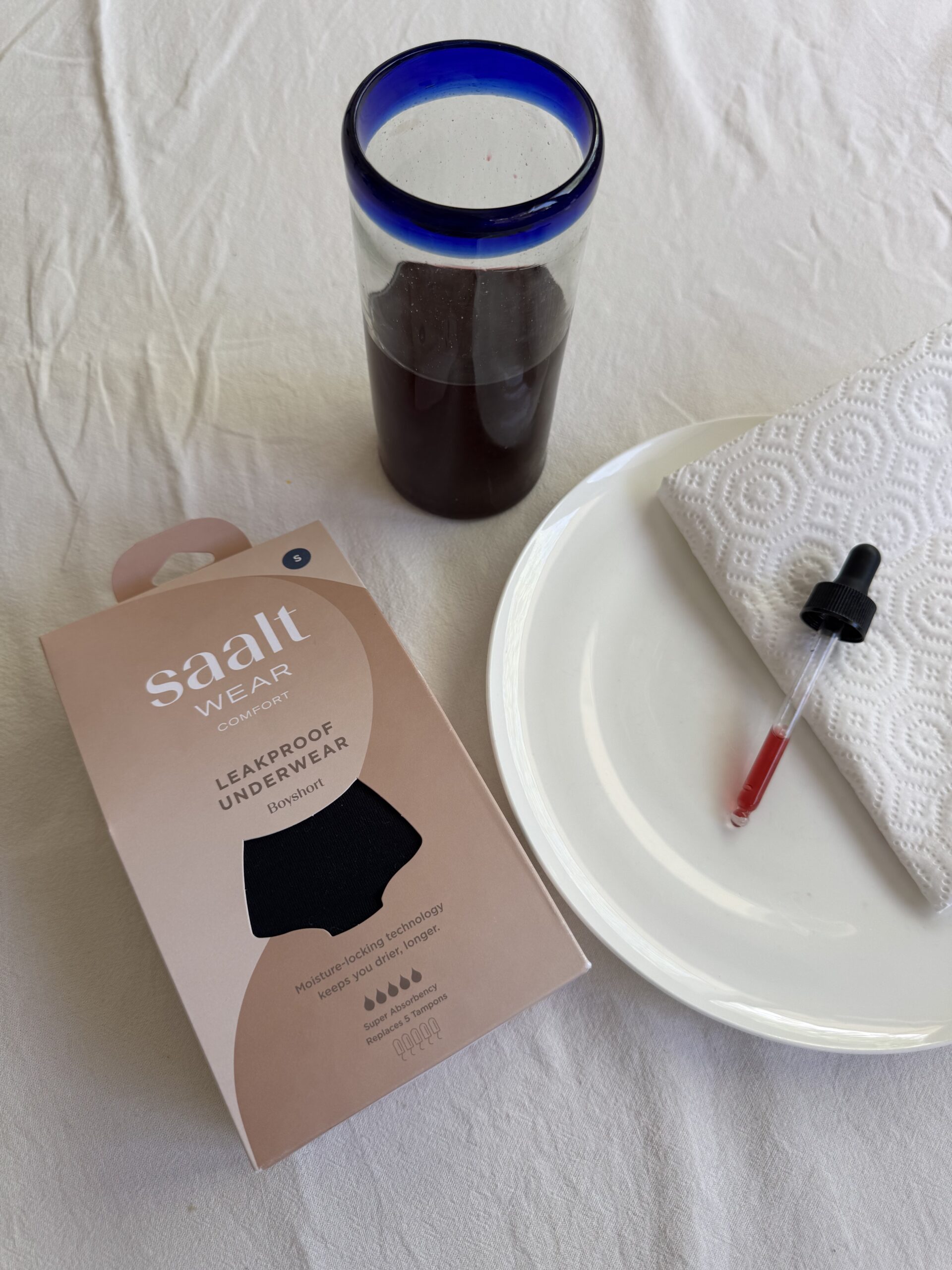 Box of leakproof underwear, glass of dark liquid, plate with a napkin, and a dropper with red liquid on a table.