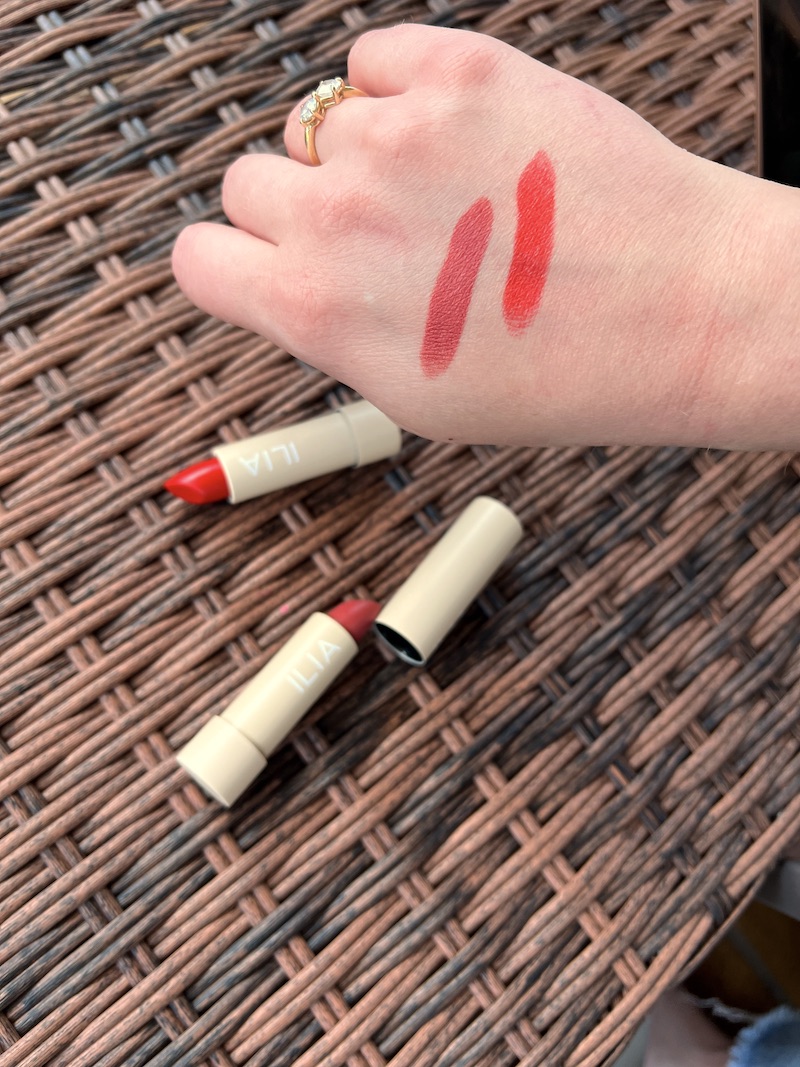 A hand with two lipstick swatches on the back is placed on a wicker surface. Two lipstick tubes with caps off are nearby, revealing the shades swatched on the hand.
