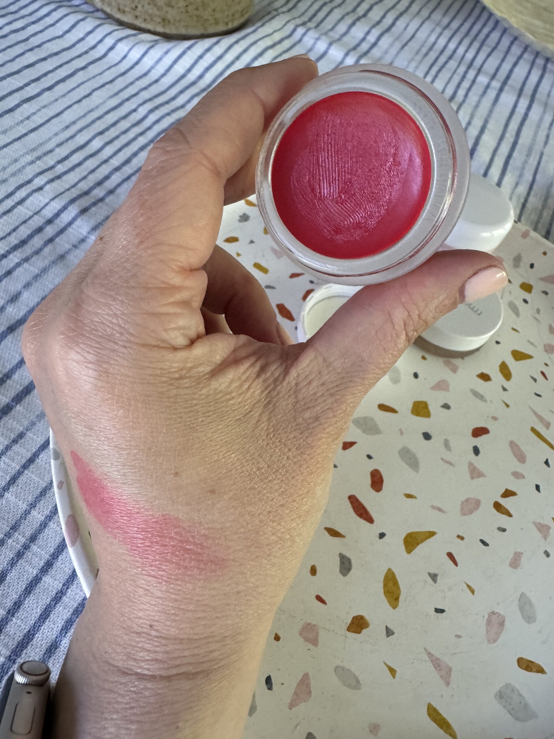 A person holds an open container of bright pink lip balm with a swatch of the same color on the back of their hand, resting on a patterned tray.