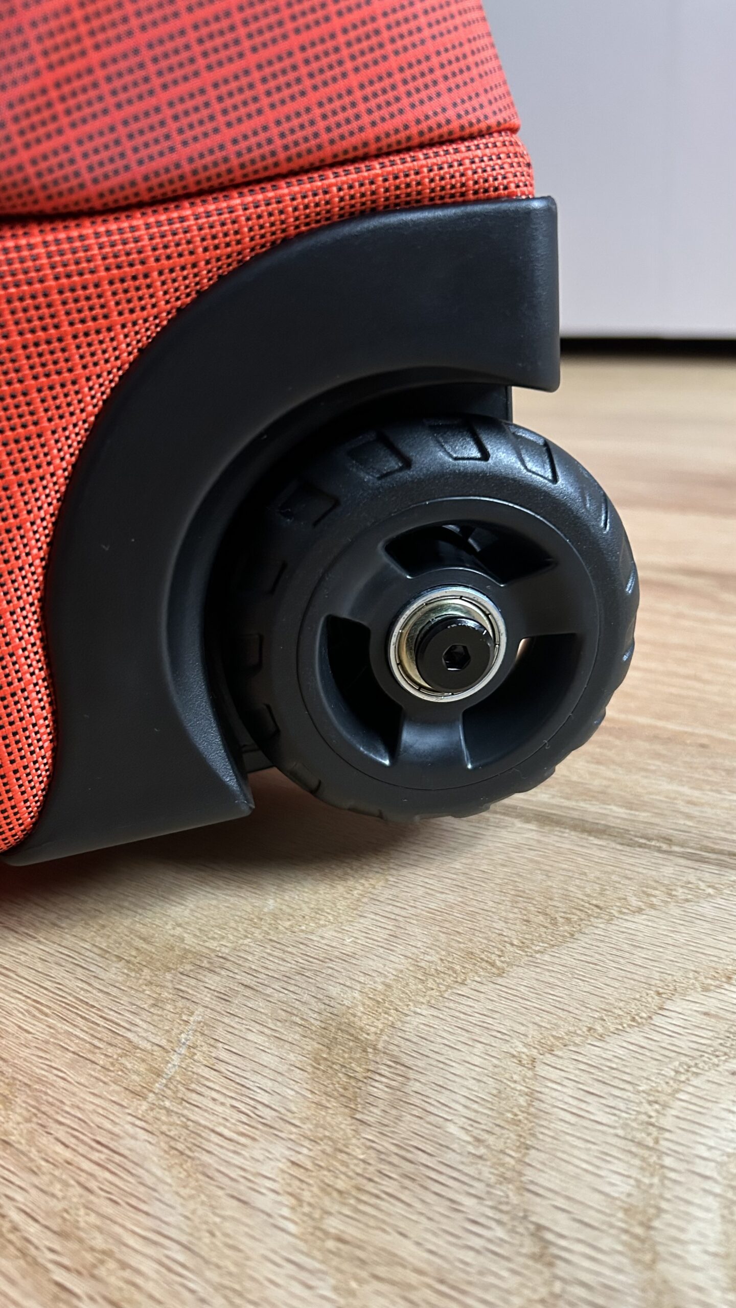 Close-up of a black plastic wheel attached to the corner of a red textured suitcase, resting on a wooden floor.