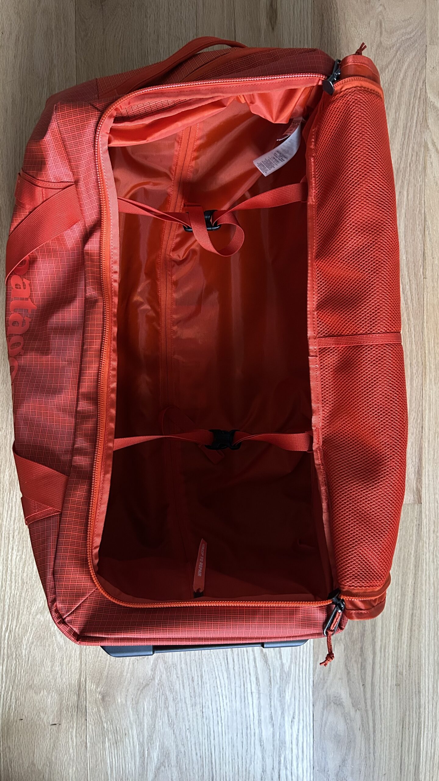 An empty red duffel bag with its main compartment open, showing two interior straps and a mesh pocket, placed on a wooden floor.