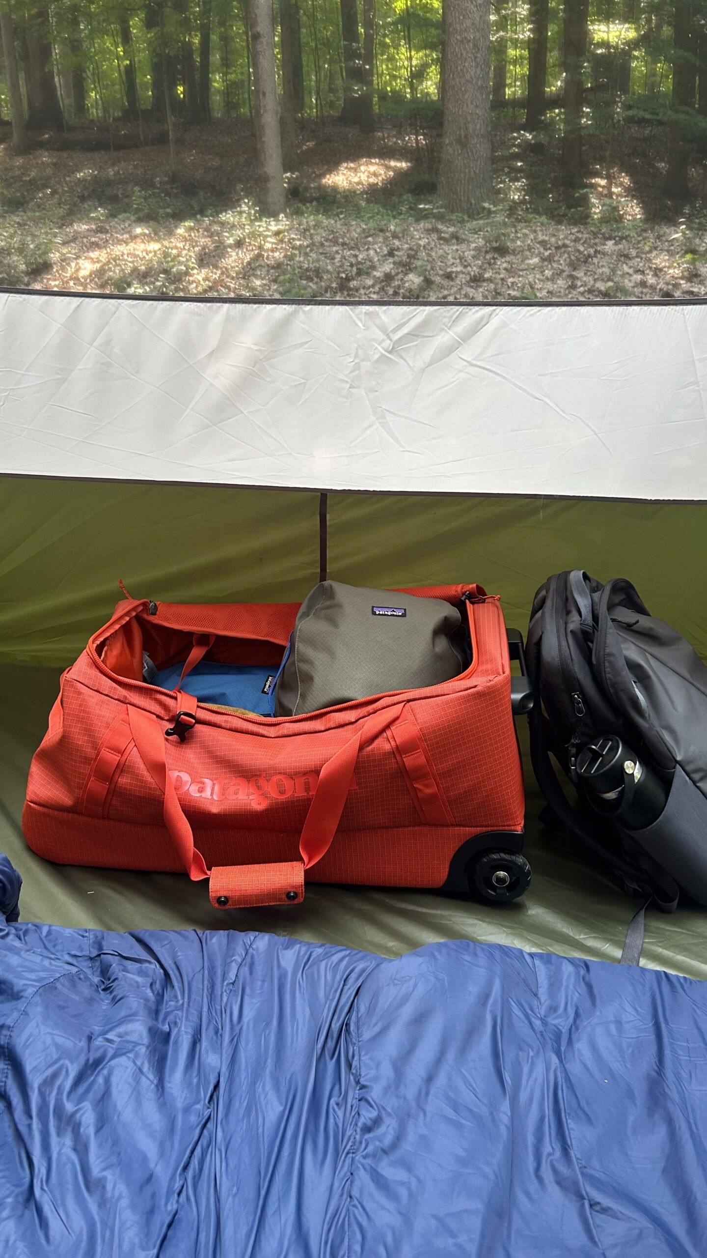 A red rolling duffel bag, a gray backpack, and a green bag are placed inside a tent with a blue sleeping bag visible in the foreground and a forest visible outside.