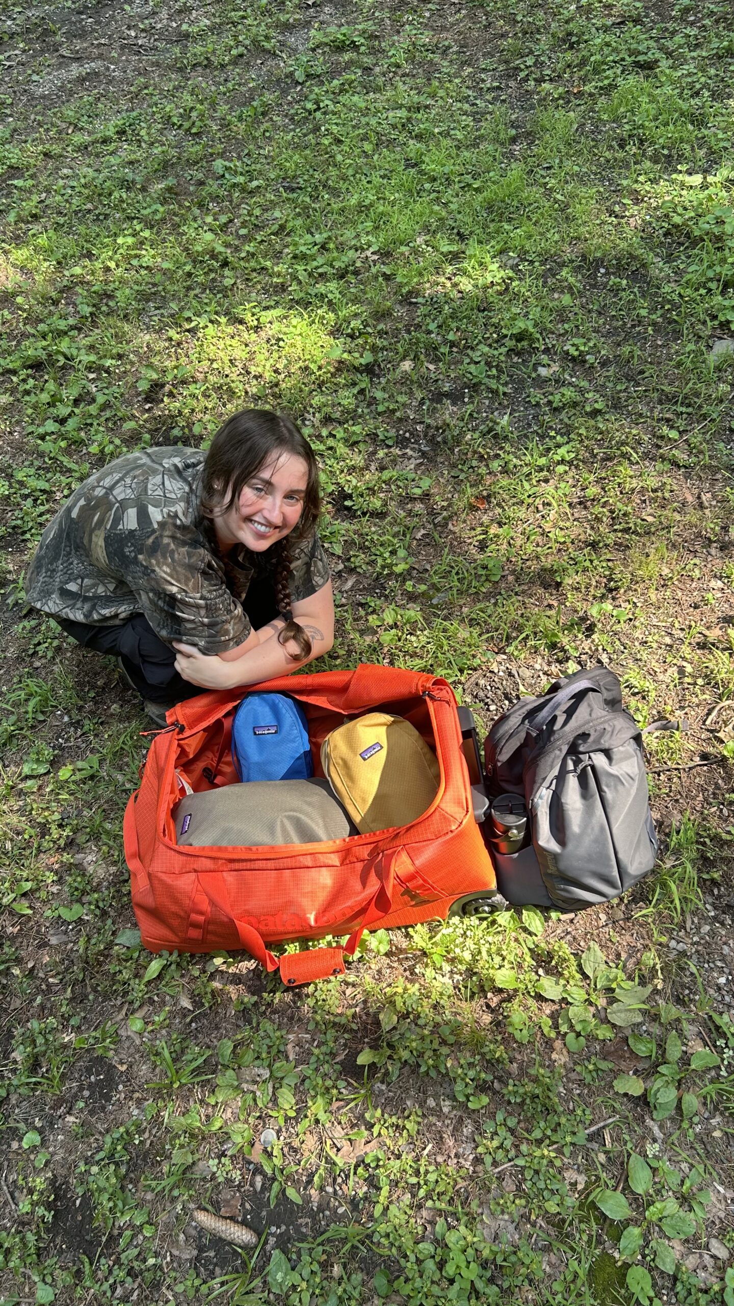 Person kneeling on grass next to an open orange duffel bag filled with colorful pouches and a gray backpack.