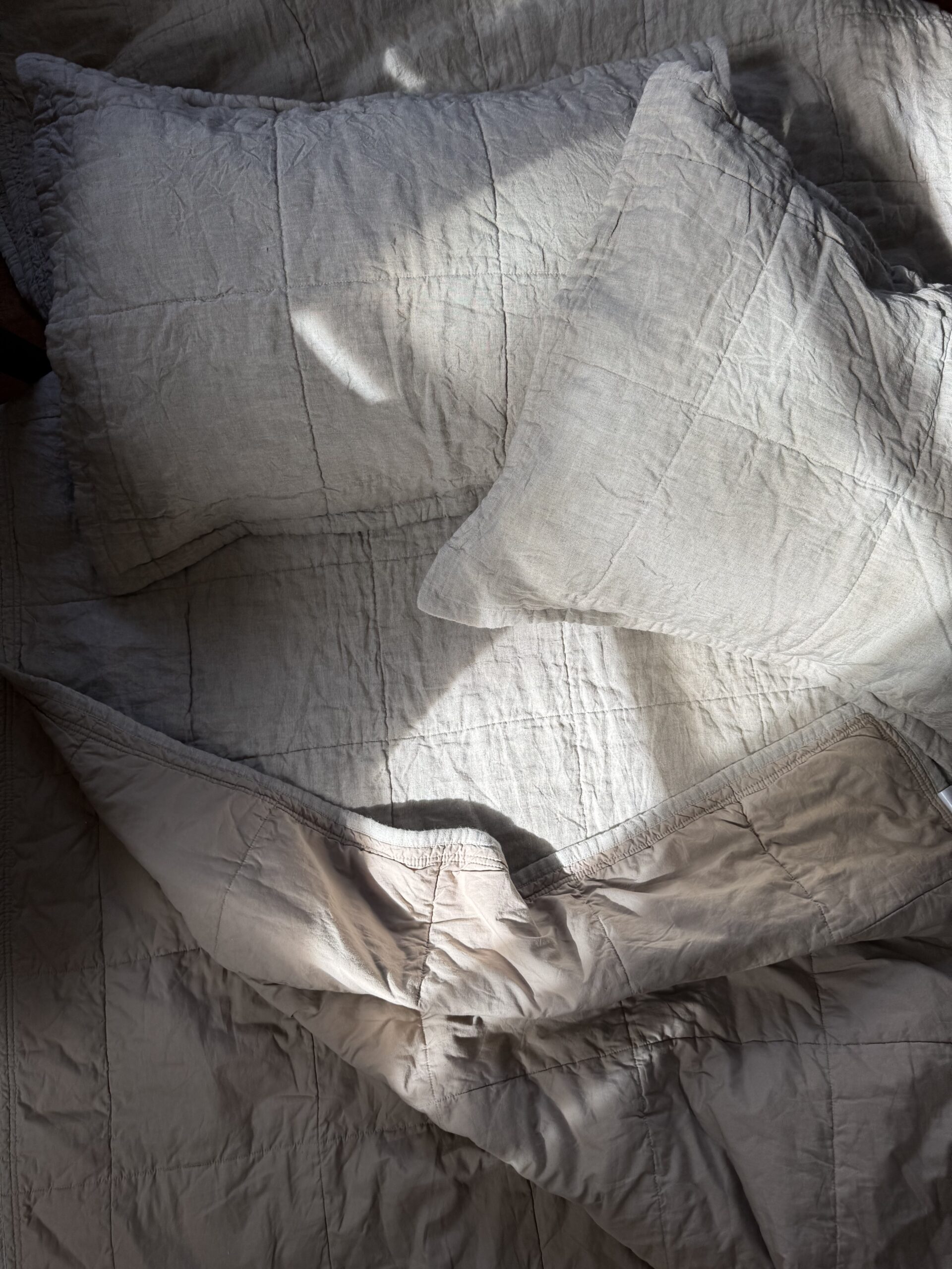 A crumpled gray bedspread and two pillows are illuminated by soft sunlight in a bedroom.