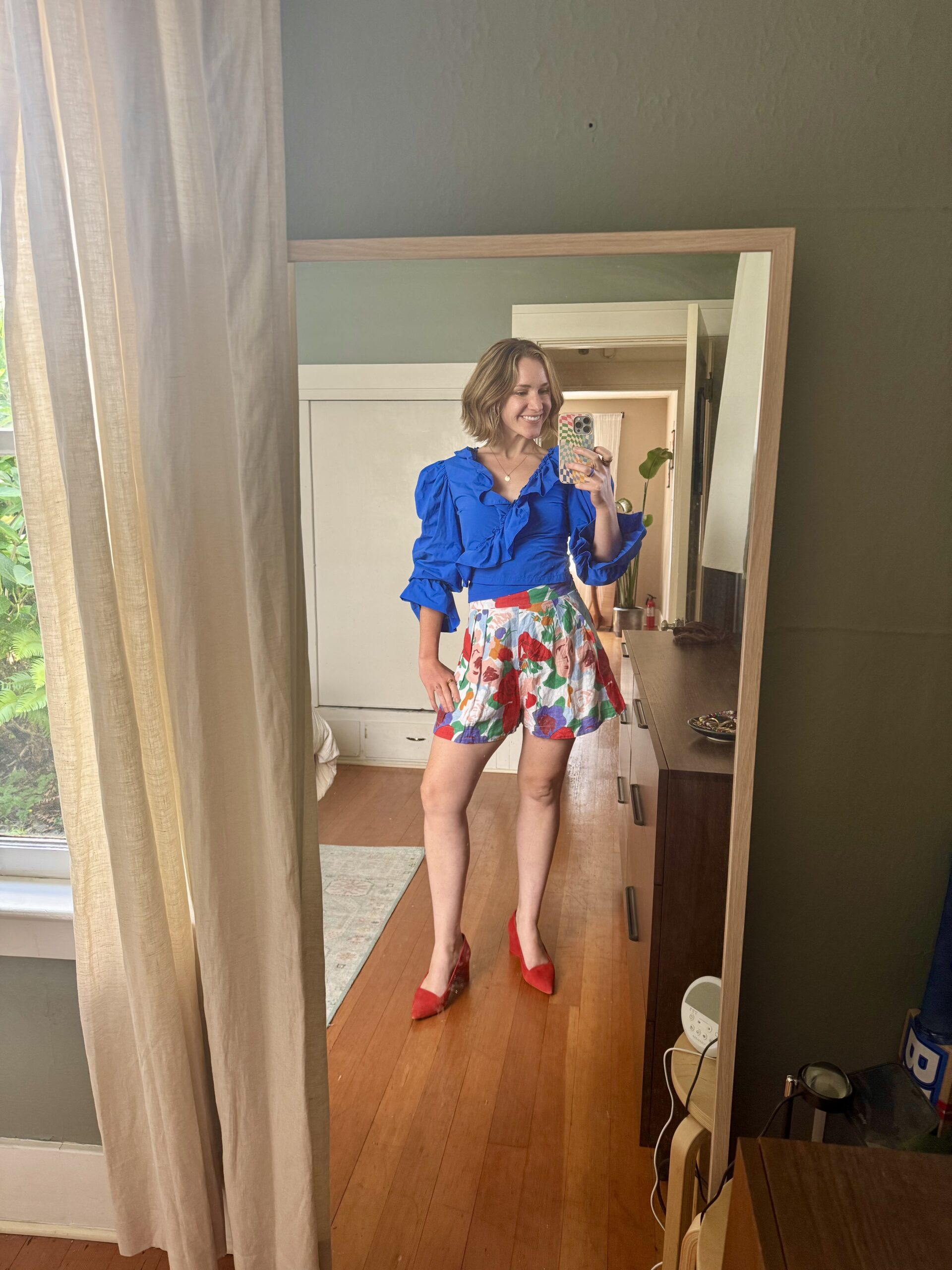 A person stands in front of a mirror, wearing a blue blouse, floral shorts, and red shoes, taking a selfie in a bedroom with wooden floors and green walls.