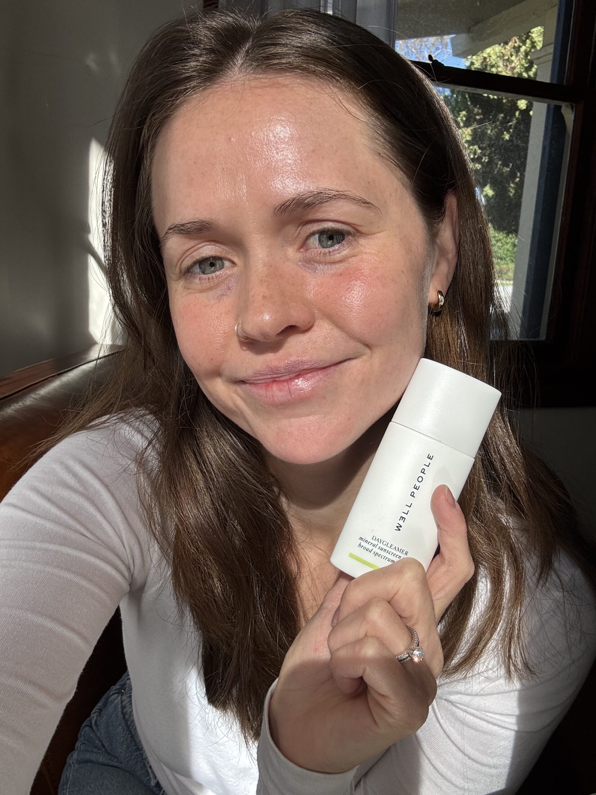 Person smiling and holding a white skincare product bottle indoors near a window.