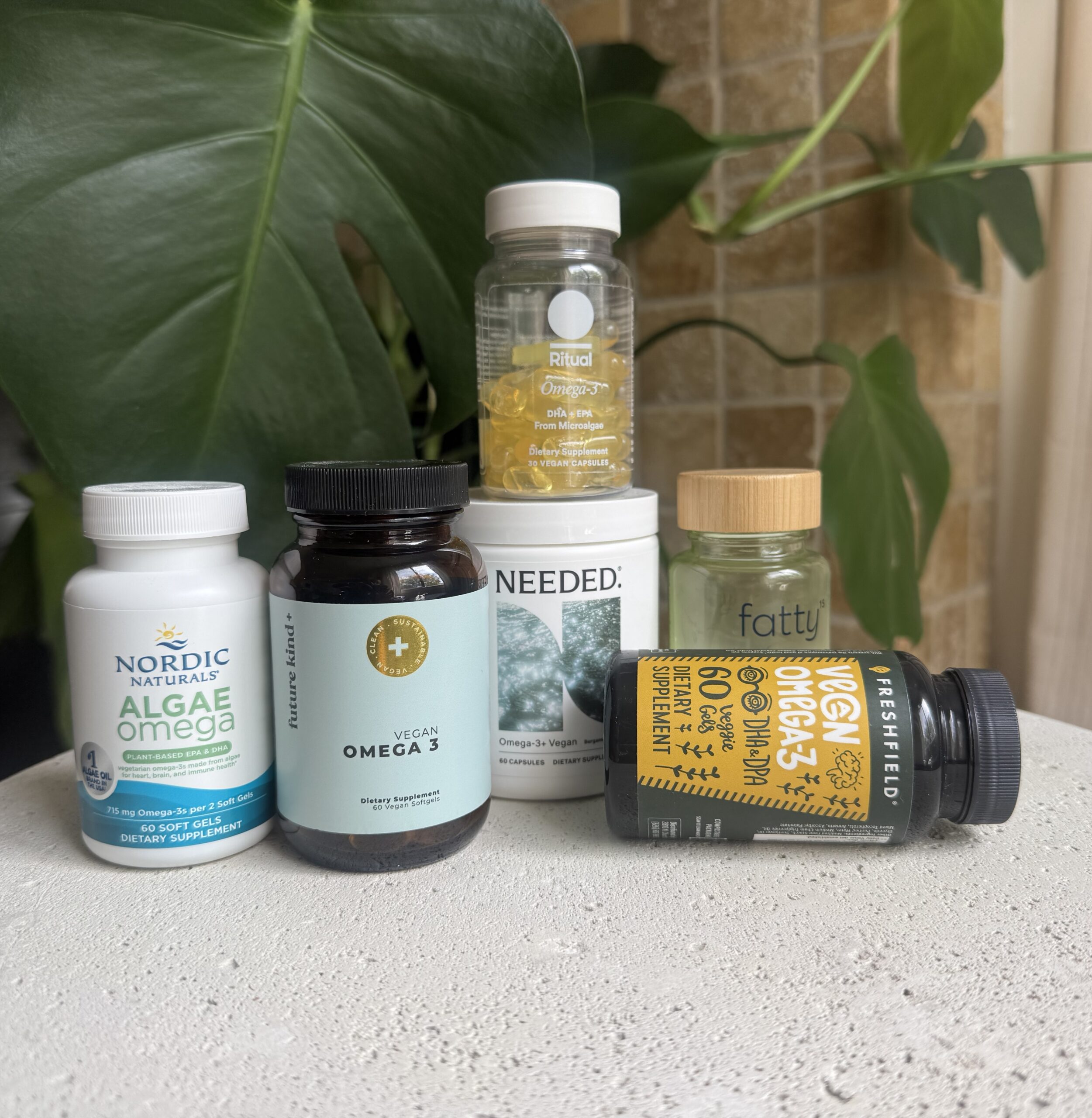 Six different omega-3 supplement bottles are arranged on a white surface in front of a large green plant.