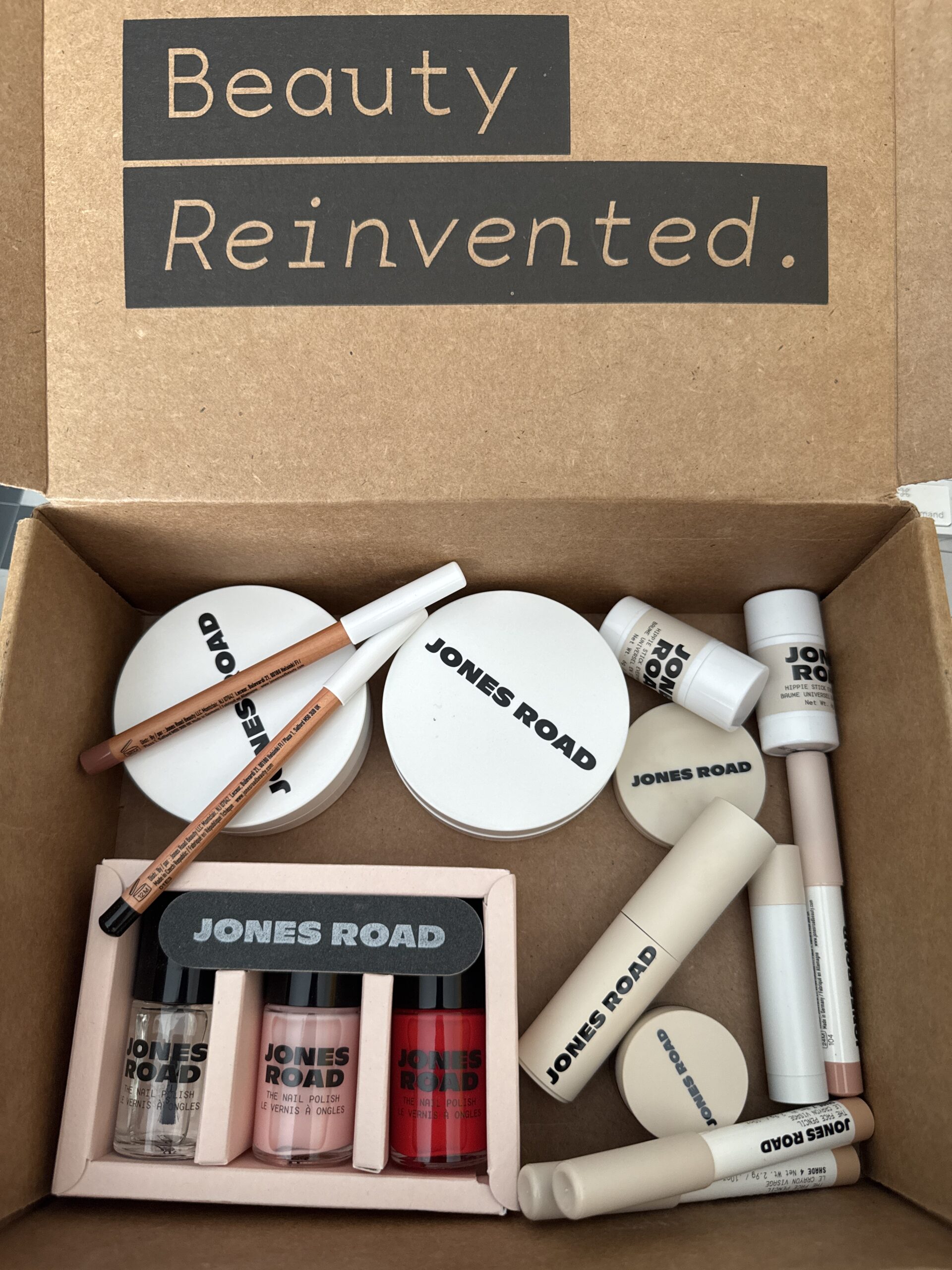 Box of Jones Road and Ilia cosmetics featuring lip glosses, creams, pencils, and brushes with "Beauty Reinvented" elegantly inscribed on the lid.