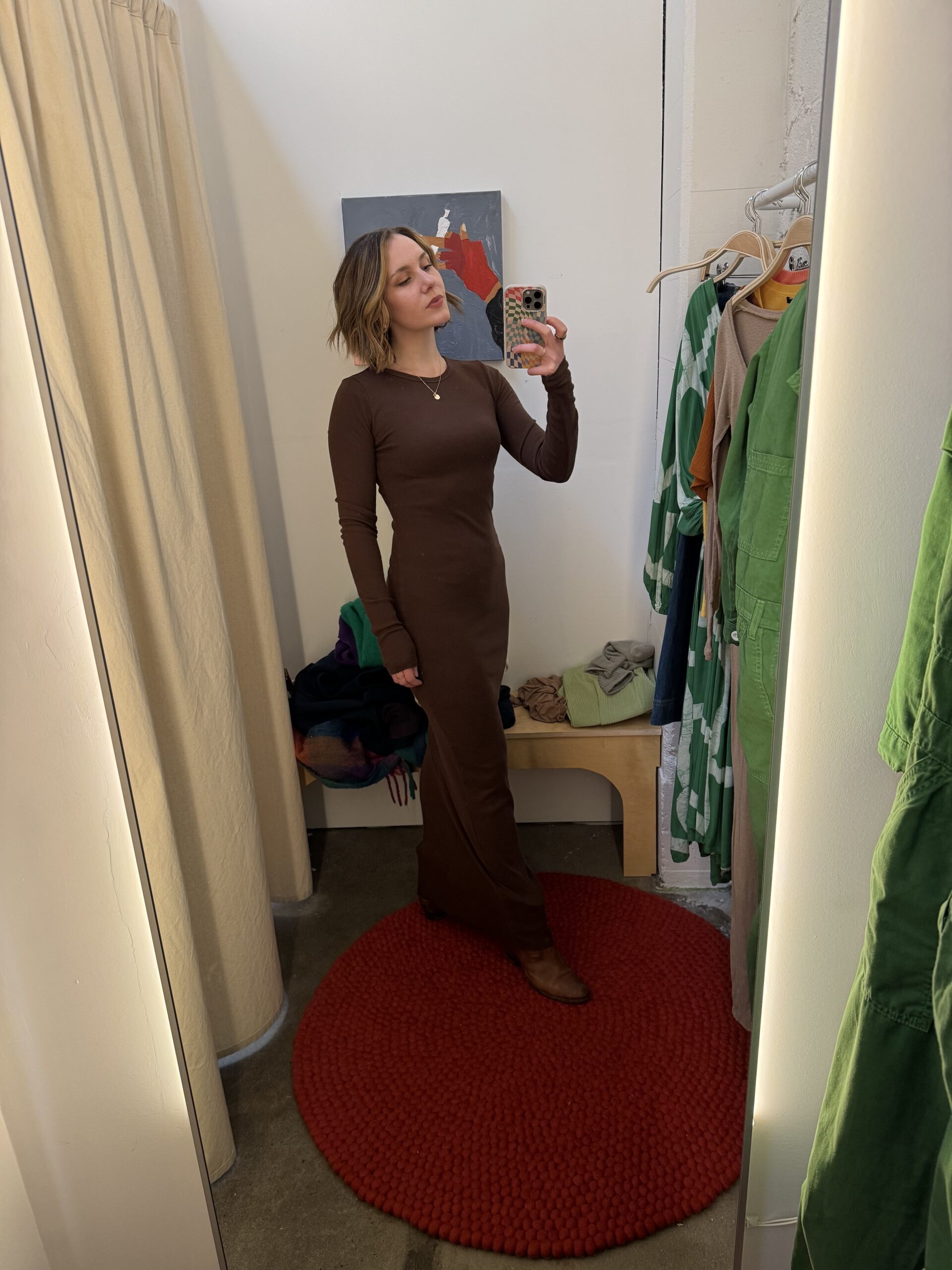 Woman takes mirror selfie in a fitting room while wearing a long brown dress, standing on a red circular rug. Clothes and a bench are visible in the background.