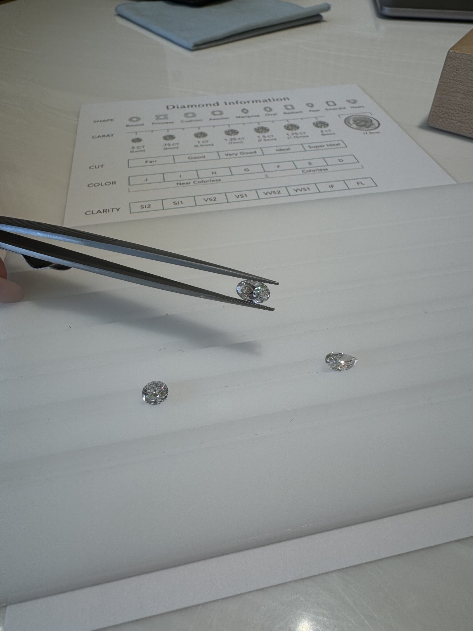 A hand holds a diamond with tweezers above a white surface with two other loose diamonds nearby; a diamond grading chart is visible in the background.