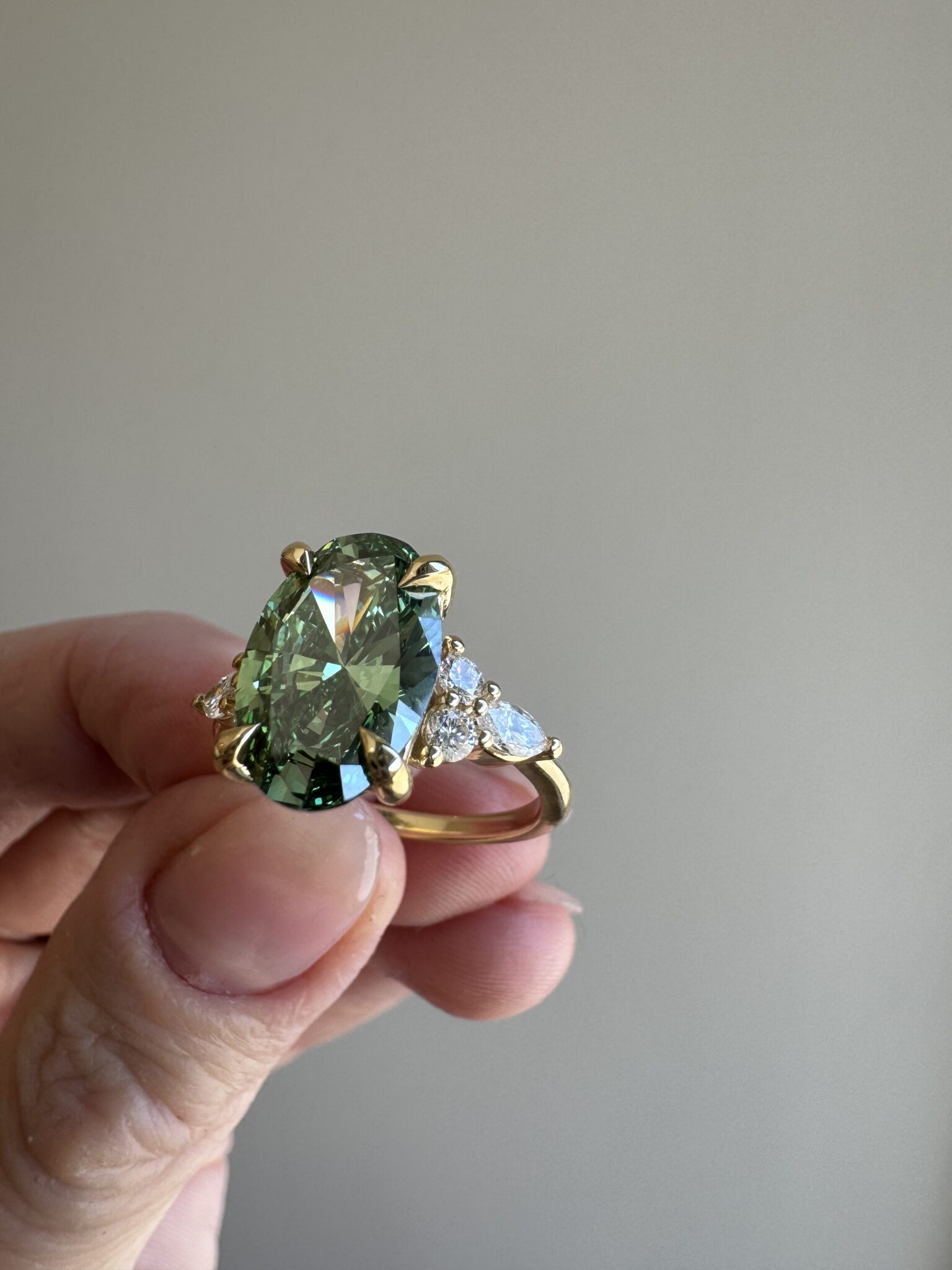 A hand holds a gold ring featuring a large green gemstone at the center, flanked by smaller clear stones, against a plain background.
