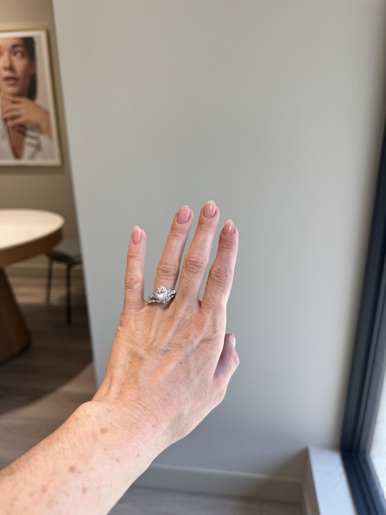 A hand with a diamond ring on the ring finger is held up in a room with light walls and a framed portrait on the wall in the background.