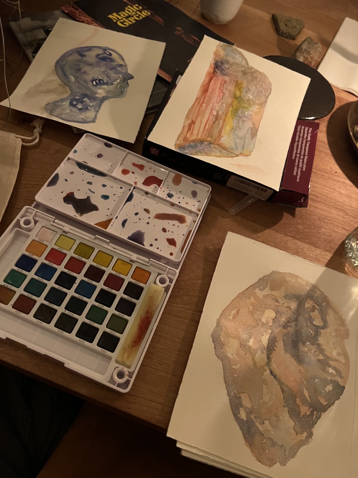 A wooden table with three watercolor paintings, a palette of paints, a brush, and books in the background. The paintings feature abstract and head-like shapes.