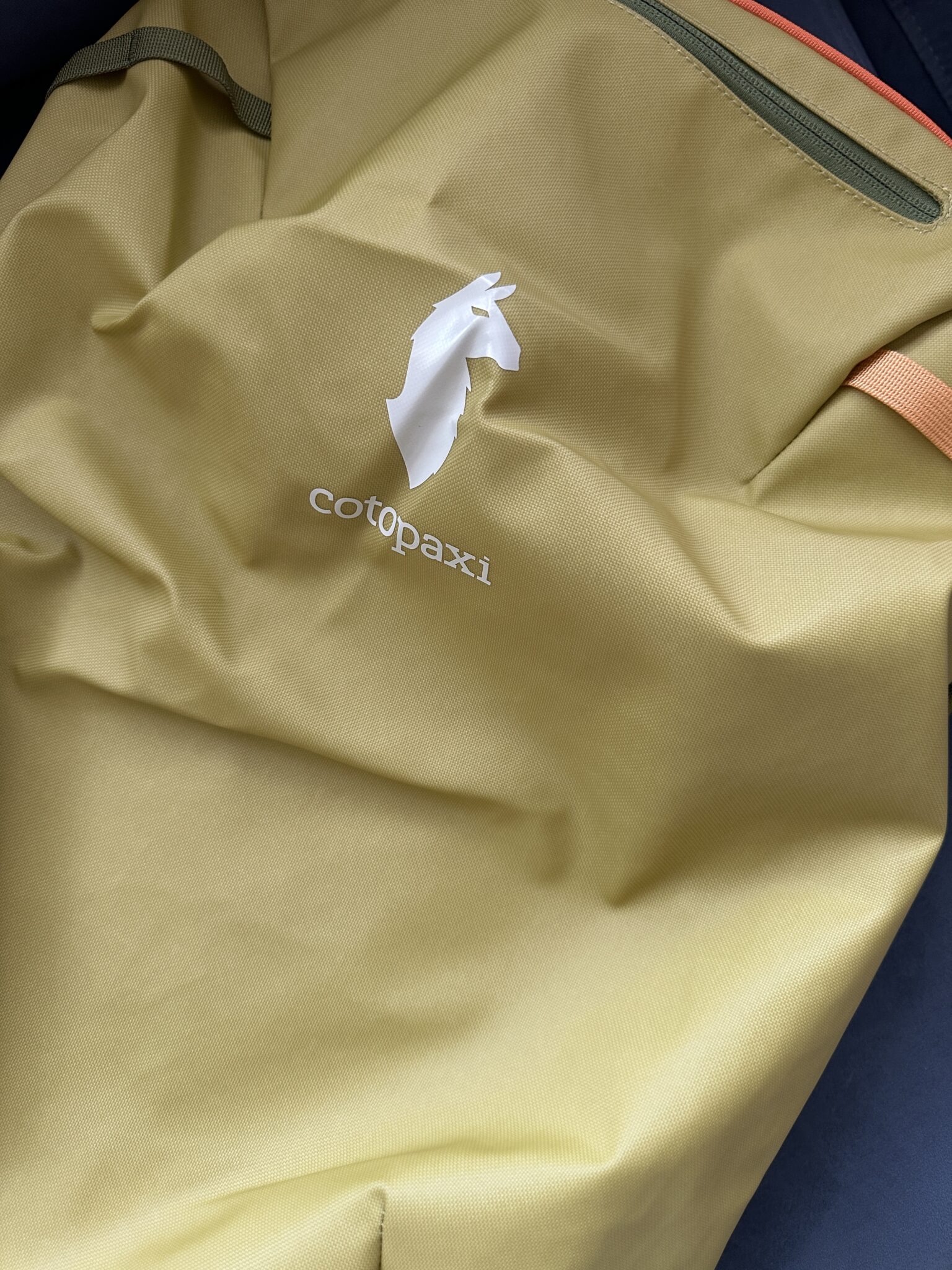 A close-up of a yellow Cotopaxi backpack with the brand’s white llama logo and name visible on the fabric.