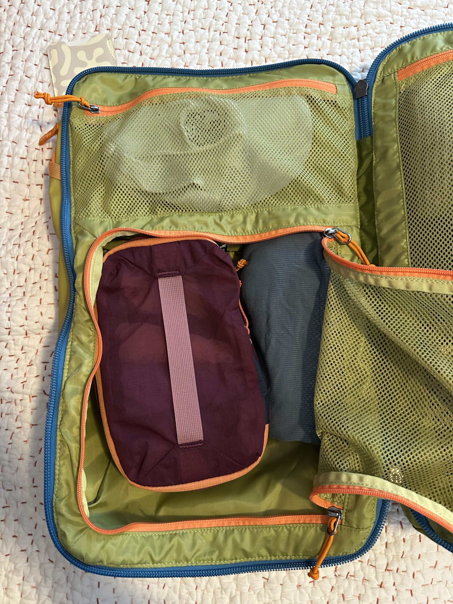 An open green travel organizer with mesh pockets contains a purple pouch with a gray strap and other packed items inside, lying on a textured white surface.