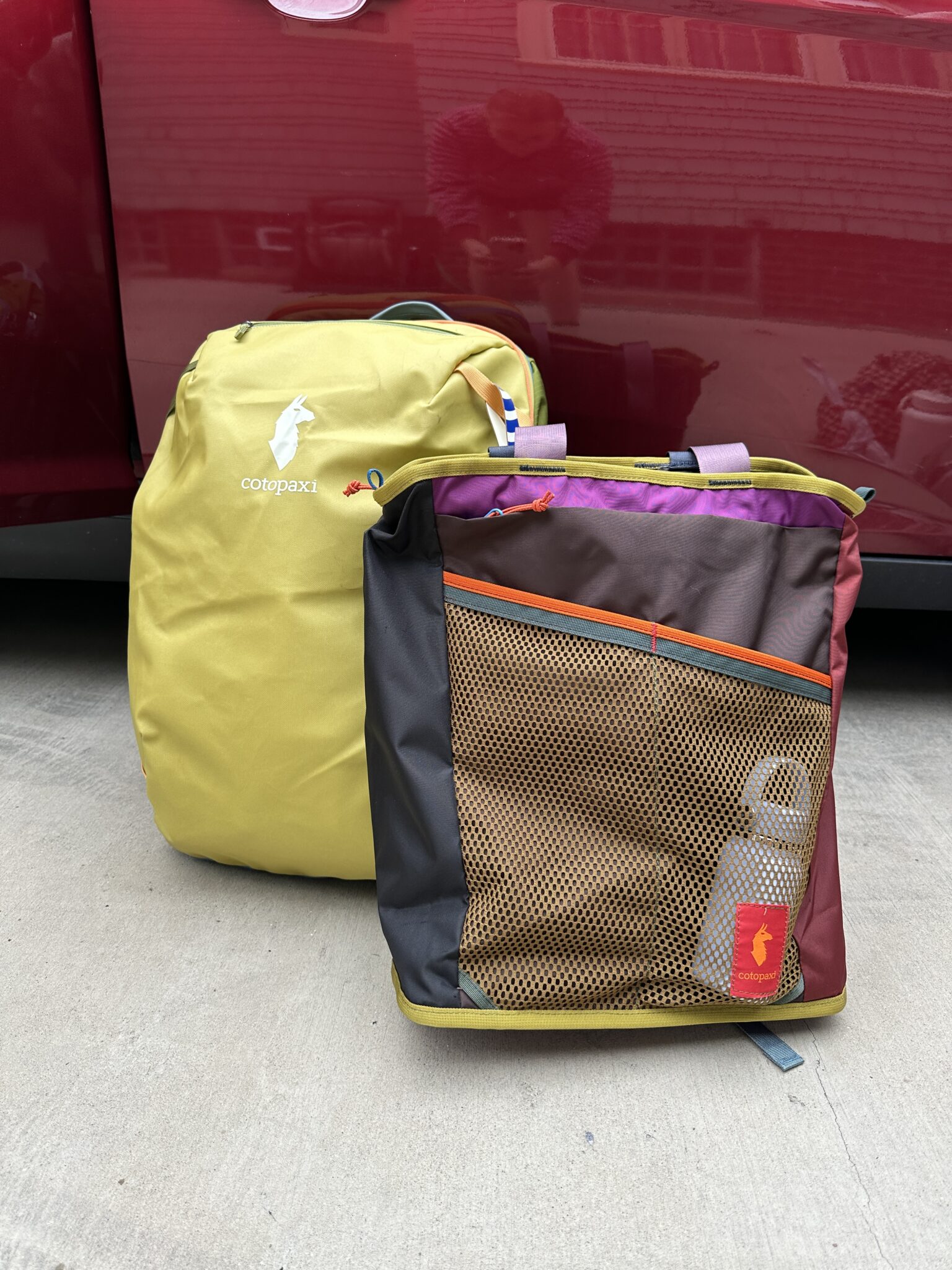 Two colorful Cotopaxi bags, one yellow and one multicolored with a mesh pocket and water bottle, are placed on concrete in front of a red vehicle.