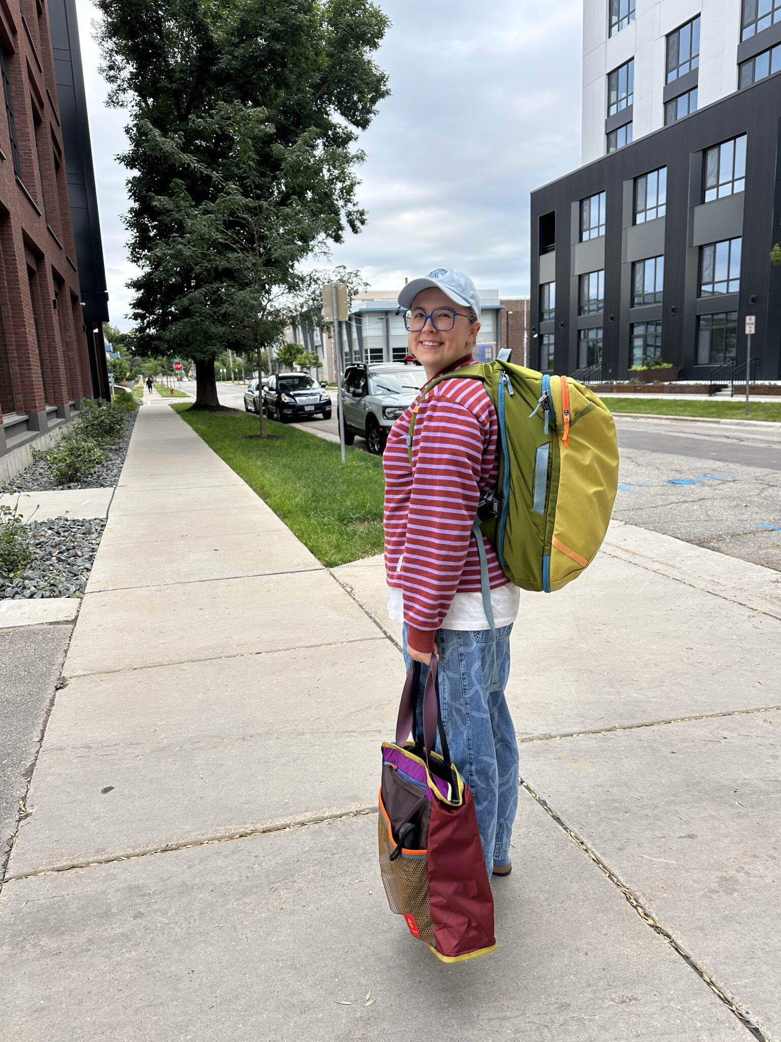 A person wearing a striped shirt, cap, and backpack stands on a city sidewalk holding a colorful tote bag, looking back and smiling.