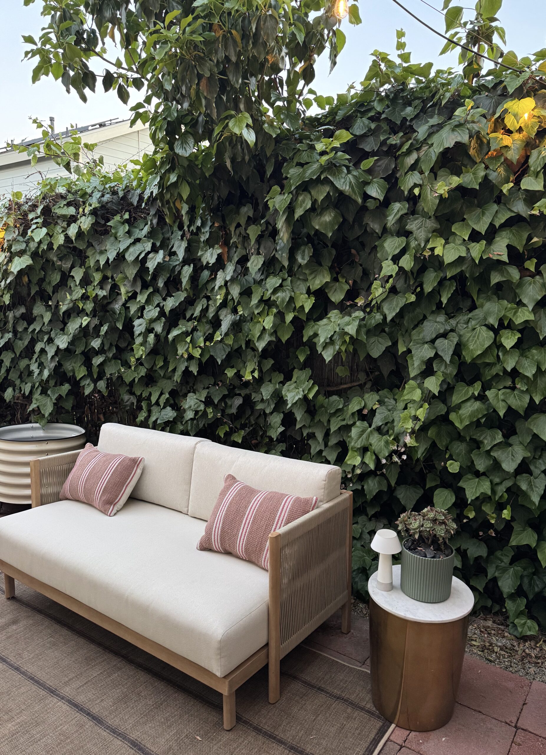 A beige outdoor sofa with striped pillows sits on a rug next to a small table with a lamp and potted plants, against a backdrop of a green ivy-covered fence.