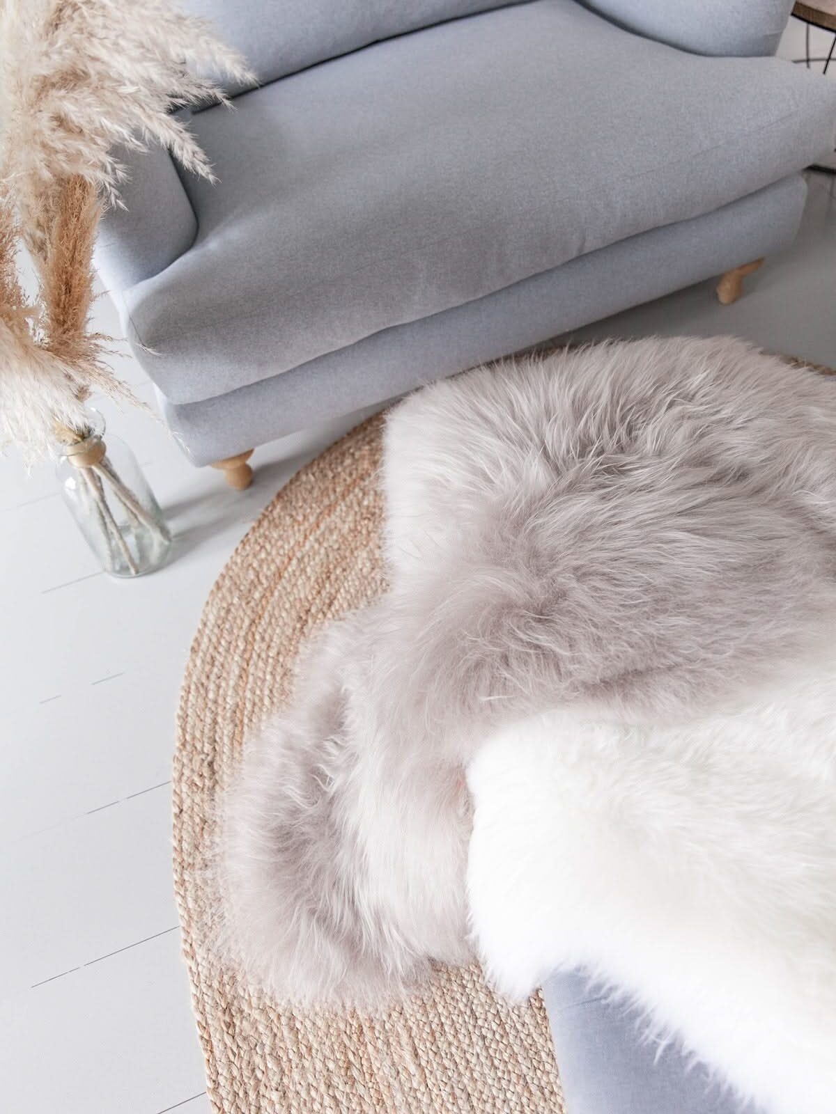 A light gray armchair sits by a round jute rug with a fluffy white and gray faux fur throw draped over it; dried pampas grass stands in a glass vase nearby.
