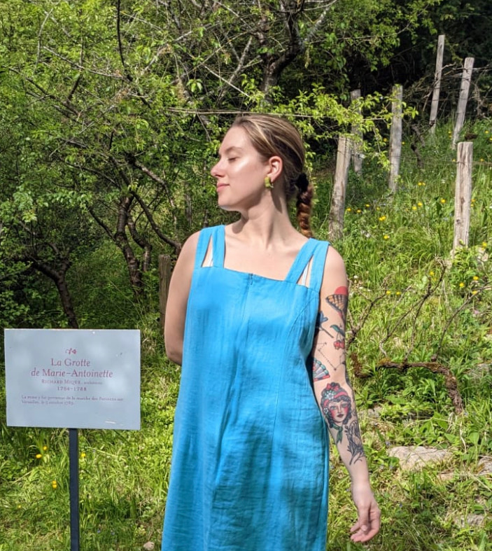A person in a blue dress stands outdoors near a grassy area with trees. There's a sign nearby in French. The person has a tattoo on one arm and is looking to the side.