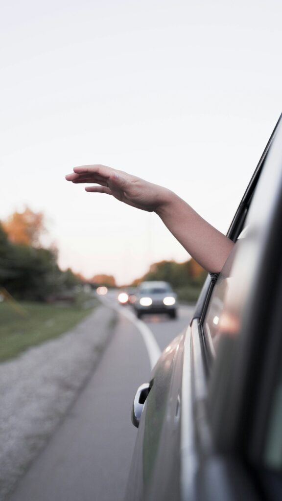 A hand extends out of a car window on a road with other vehicles in the background during daylight.