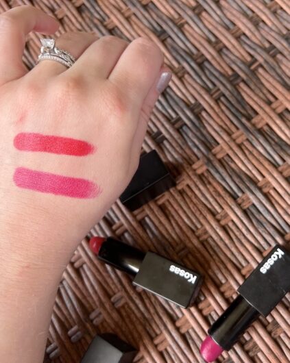 A hand with two swatches of red lipstick on the back. Four Kosas-brand lipsticks lie on a woven wicker surface. The hand is wearing a diamond ring.