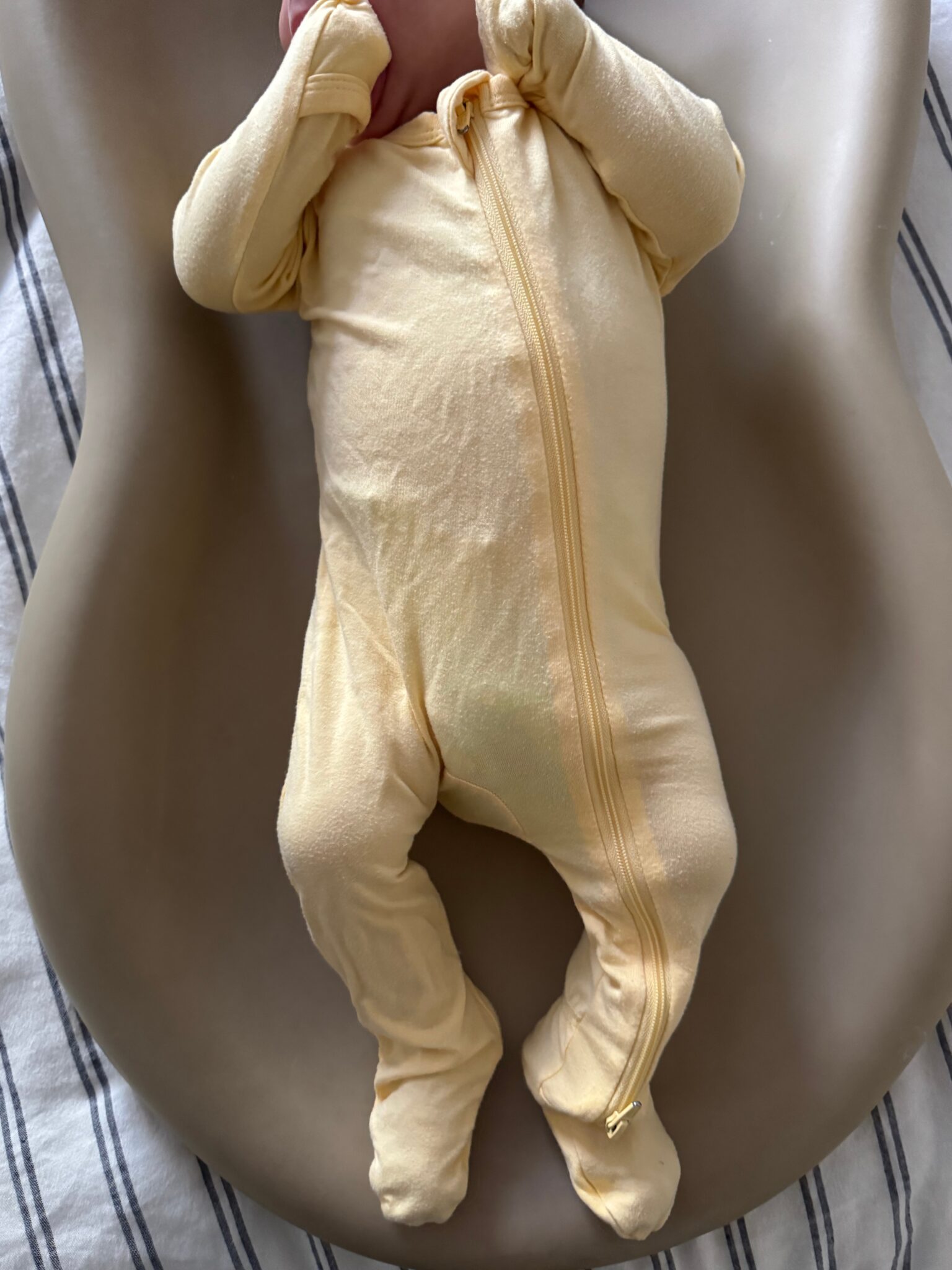 A baby in a yellow onesie with a zipper lies on a beige contoured pad, with arms raised near their head and feet covered, on a striped fabric surface.