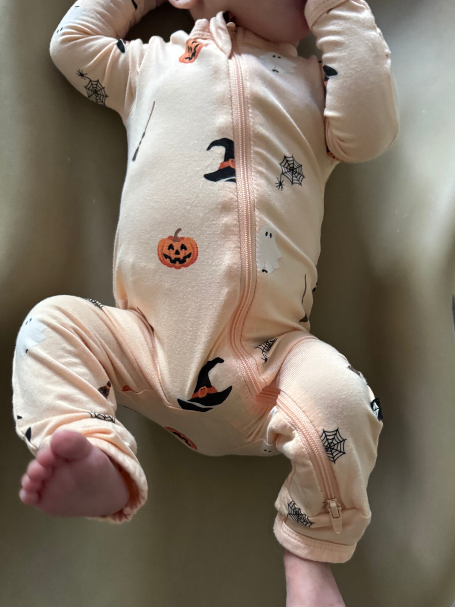 A baby in a light peach Halloween-themed onesie with pumpkins, witch hats, ghosts, and spiderwebs lies on a beige surface.