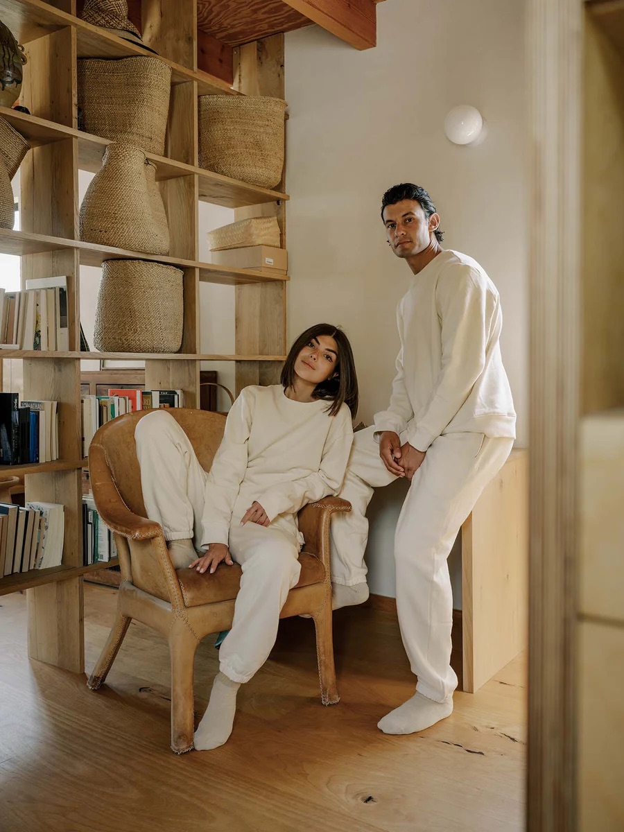 Two people dressed in cream-colored loungewear pose in a cozy, minimalist room with wooden shelves, woven baskets, books, and natural lighting—perfect inspiration for your next Labor Day sale refresh.