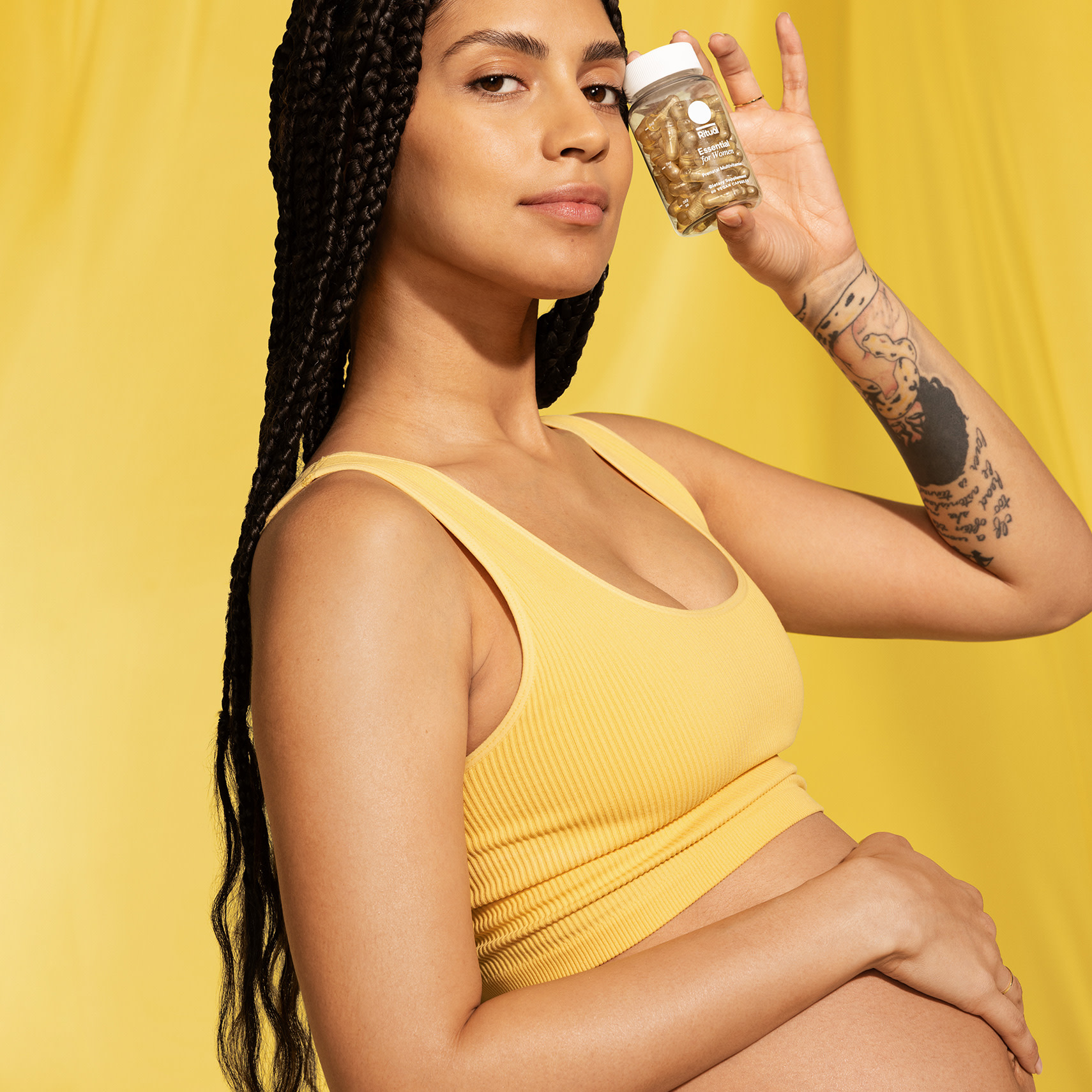 A pregnant woman in a yellow top holds a jar of capsules near her face, one hand on her belly, against a yellow background—perfect for highlighting your Labor Day sale.
