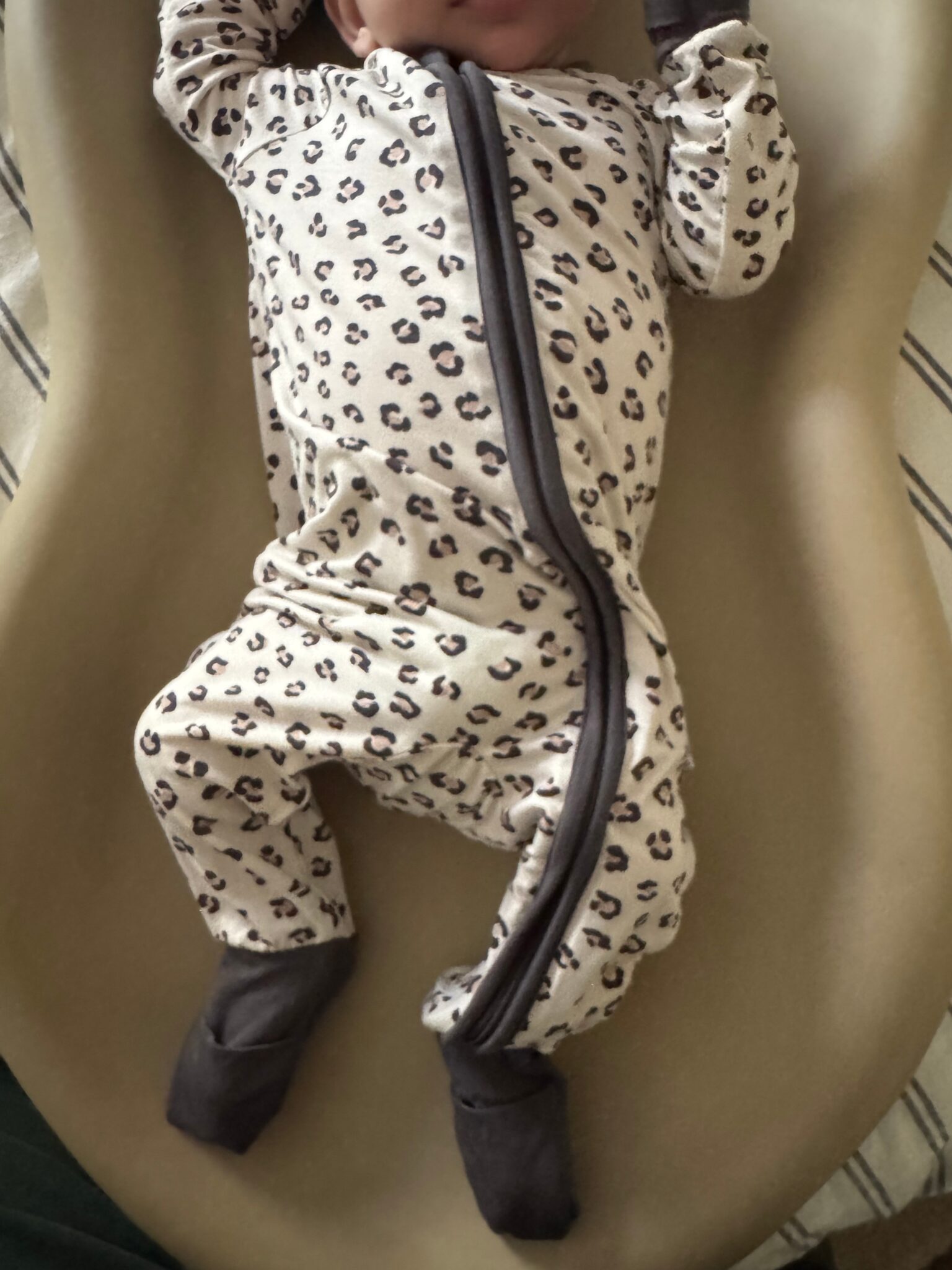 Baby lying on a beige surface wearing a cream-colored onesie with a cheetah print pattern and dark gray socks.