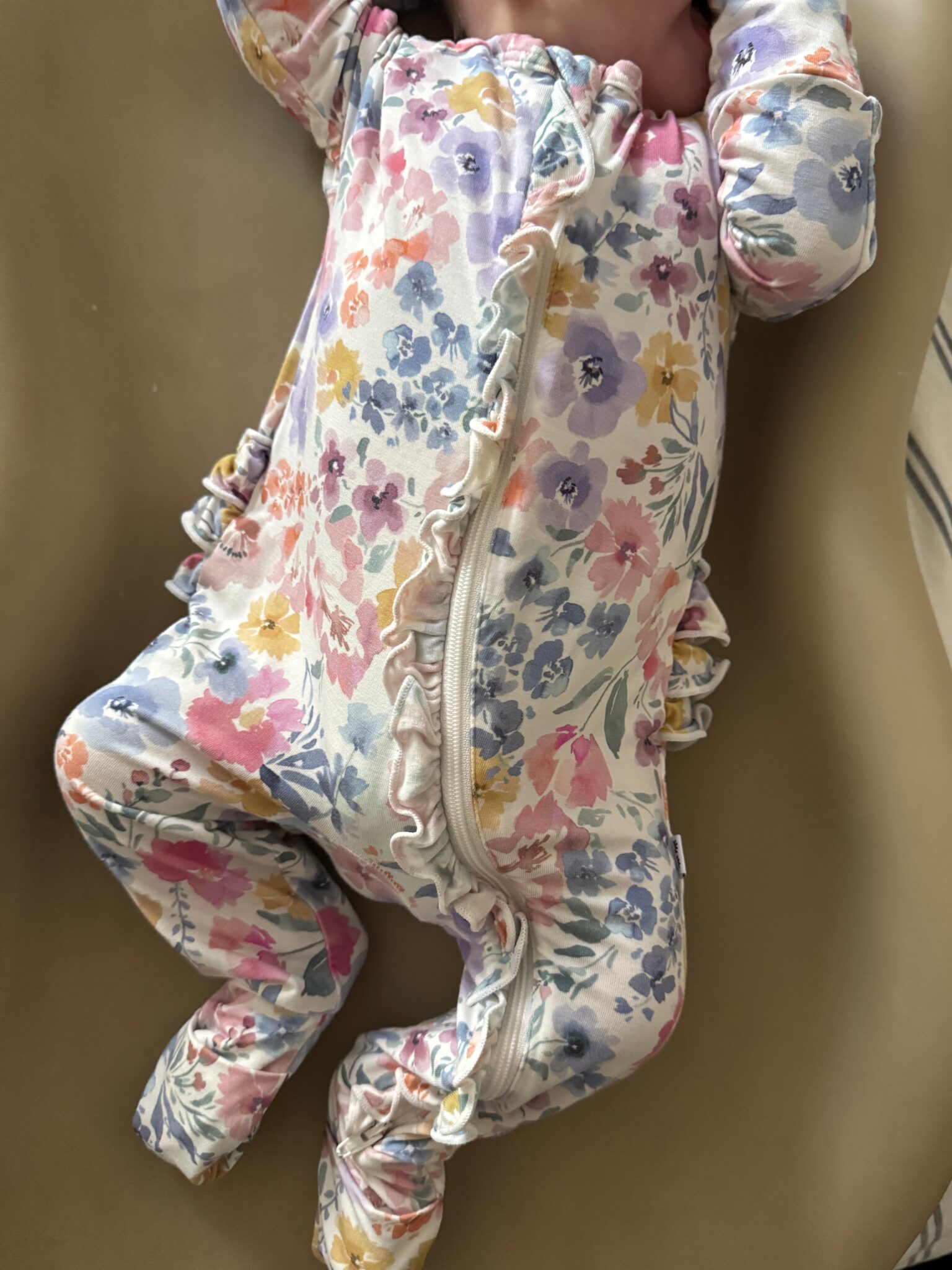 An infant wearing a floral-patterned footed onesie with ruffle details lies on a beige surface.